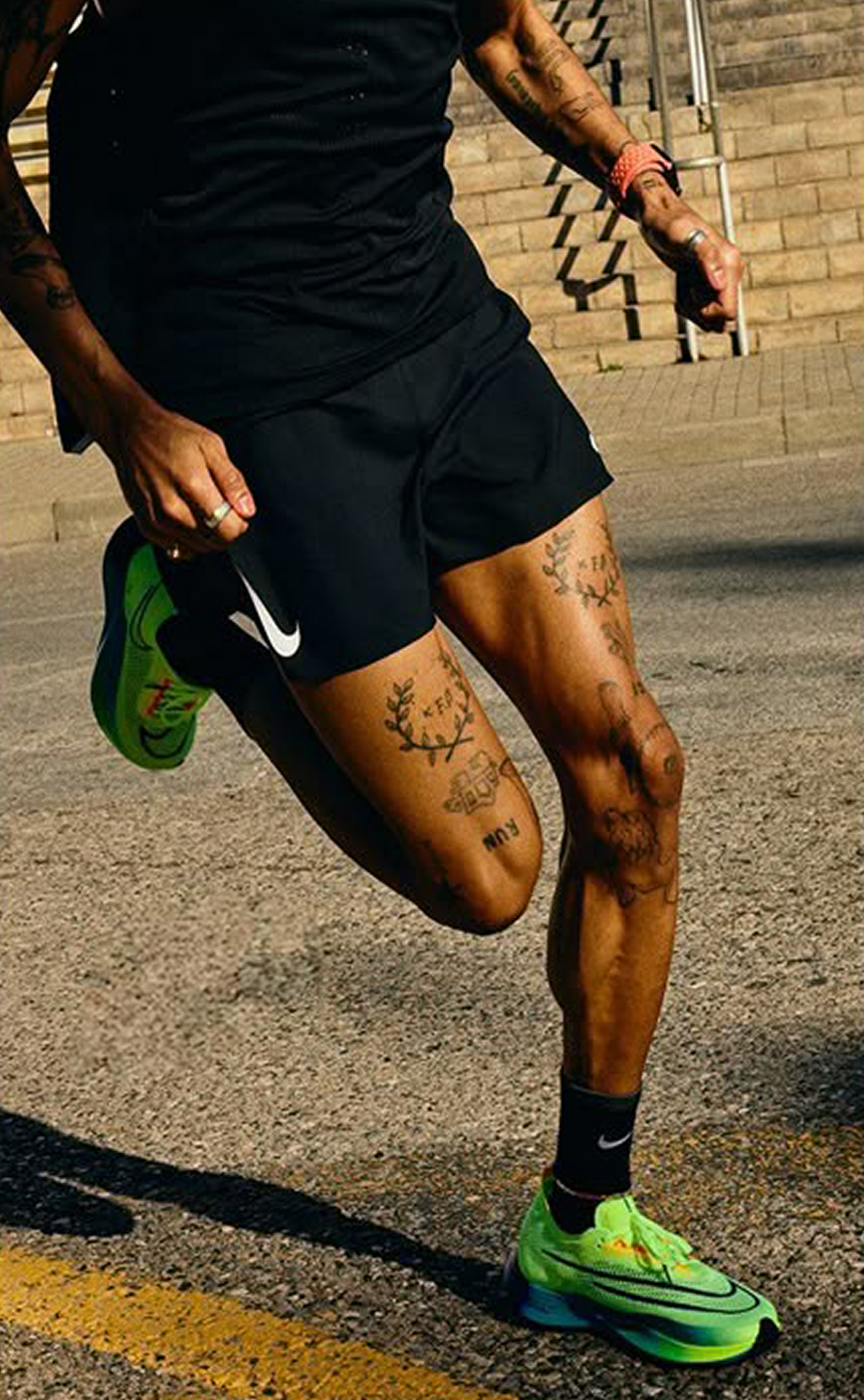 Runner in black shorts and shirt, neon green shoes, tattooed legs, running on asphalt.