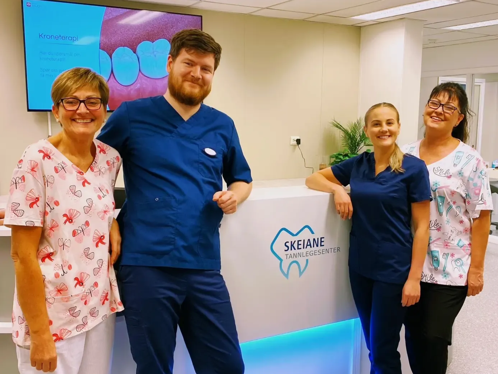 Seks kvinner i skrubber smiler i lyse omgivelser innendørs.