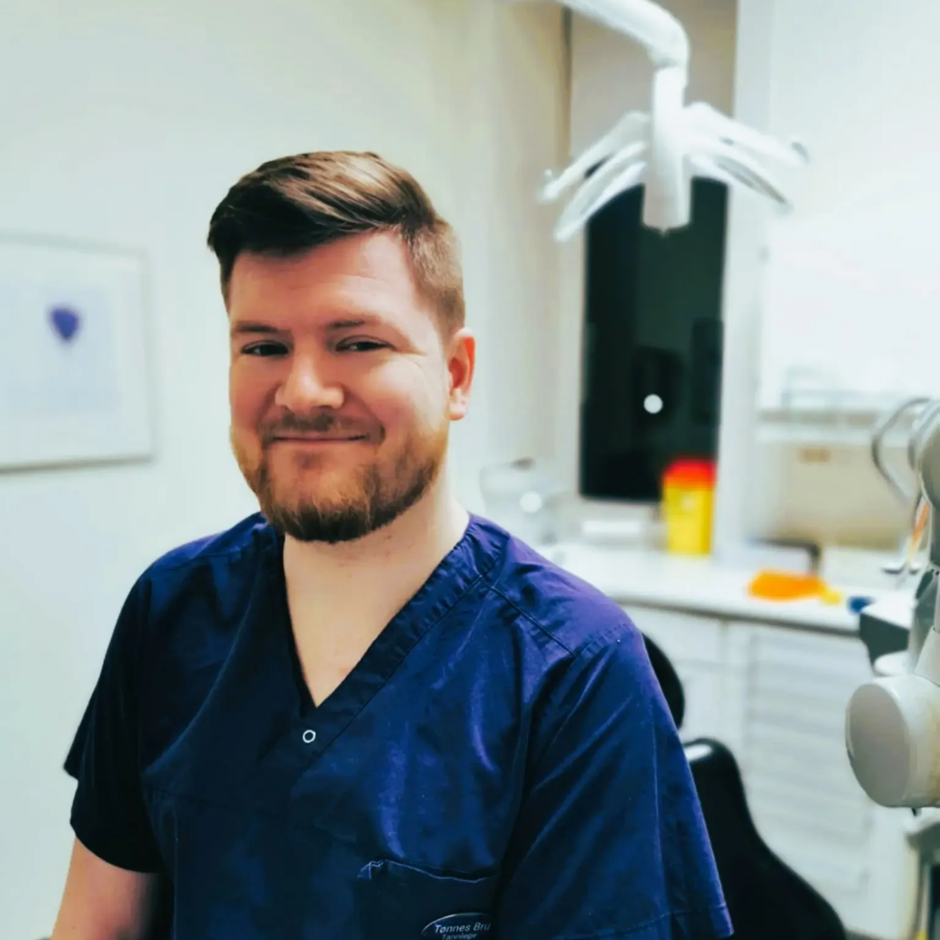 Tannlege Christian med briller og skjegg, iført blå skjorte og medisinsk maske, smiler til kameraet.