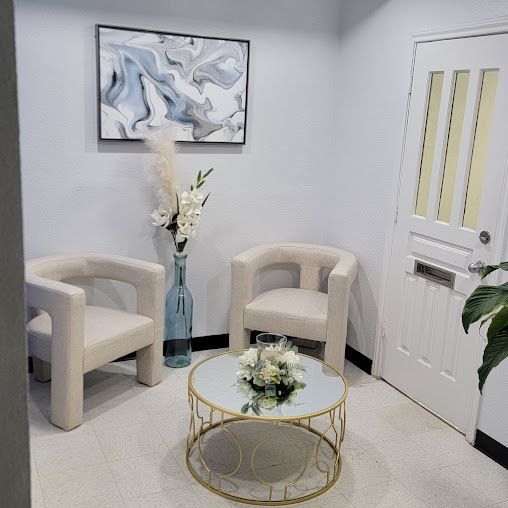 Waiting room with two beige chairs, a gold table, vase of flowers, and artwork. White walls and door.