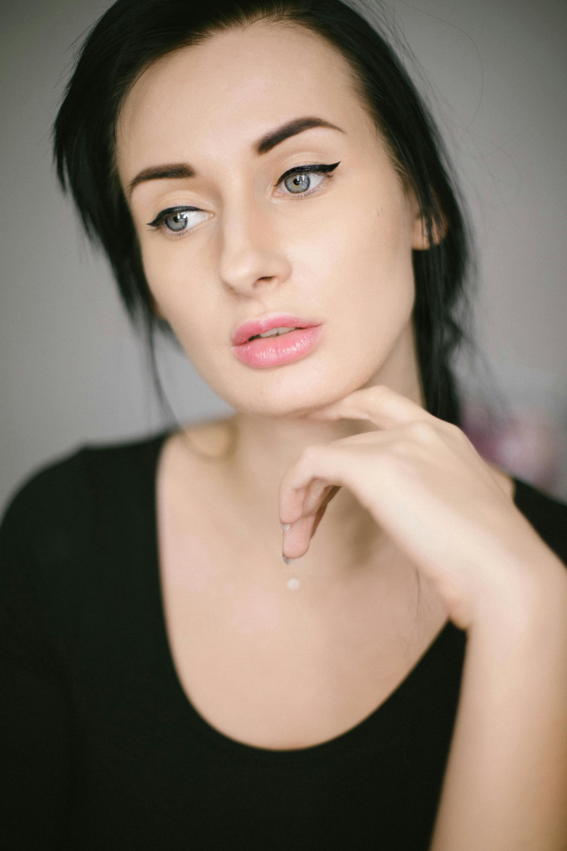 Woman in black top with winged eyeliner, hand on chin, looking away thoughtfully.