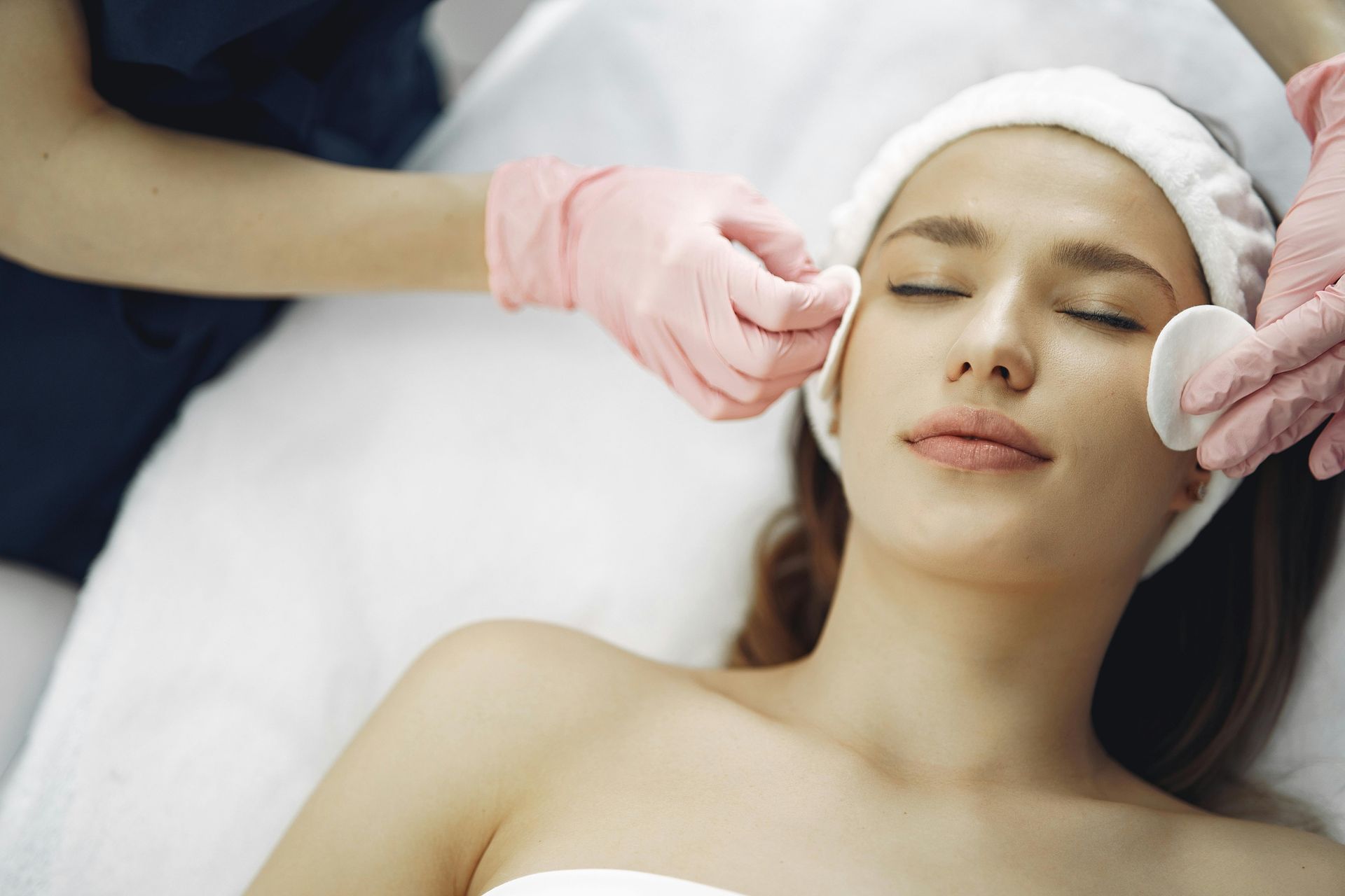 Woman receiving facial treatment; cosmetologist cleaning face with cotton pads, pink gloves.