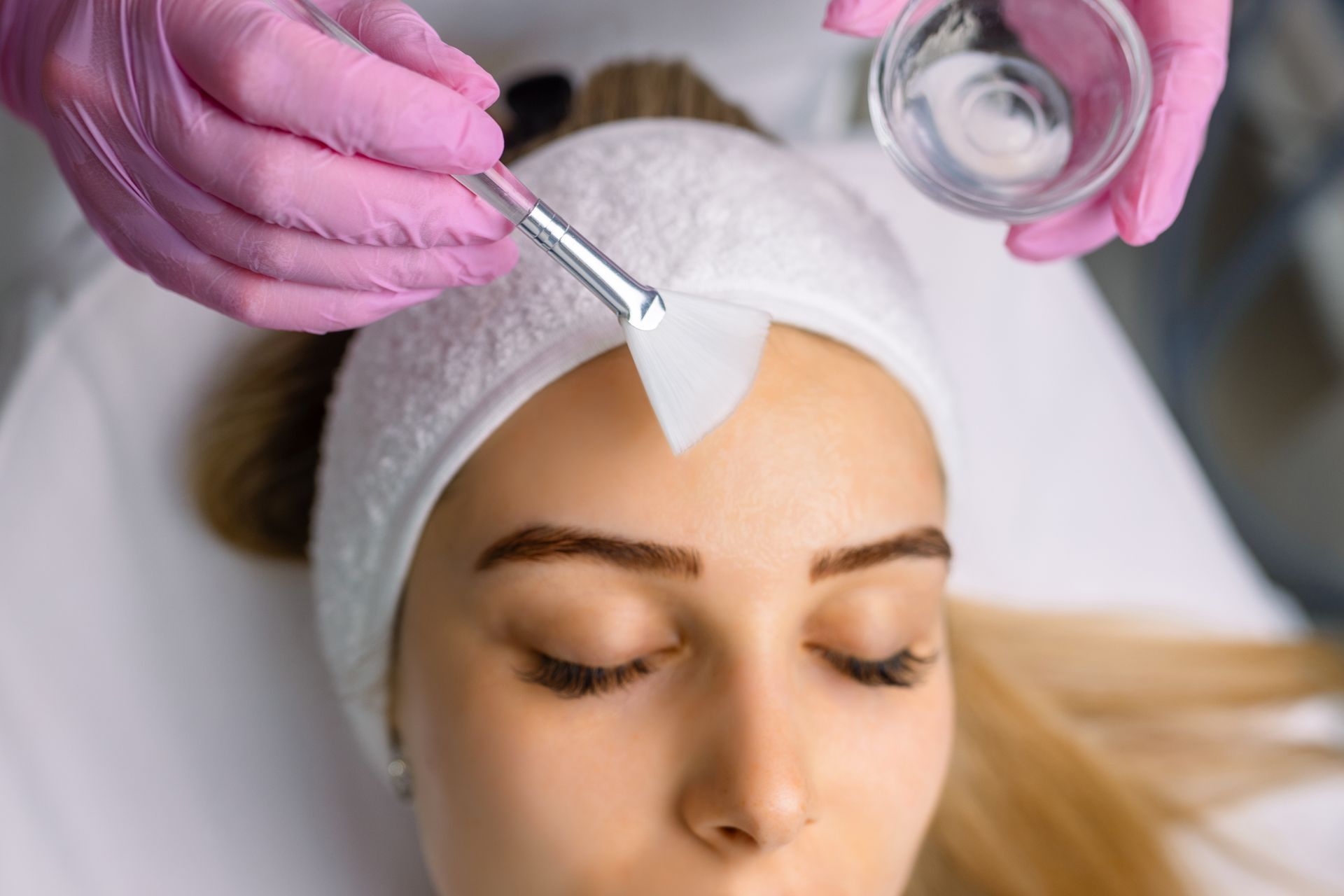 Beautician applying chemical peel to client's forehead with a brush; pink gloves, close-up.