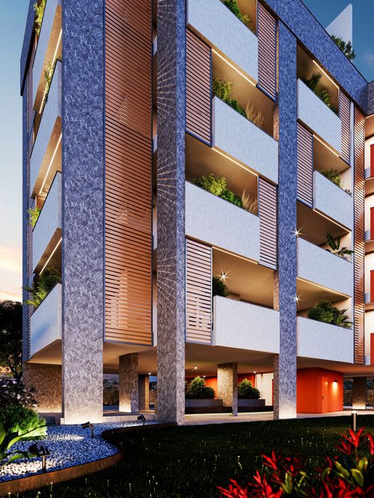 Esterno di un moderno edificio residenziale con balconi in mattoni e bianchi al crepuscolo, illuminato da luci calde e con la vegetazione sottostante.