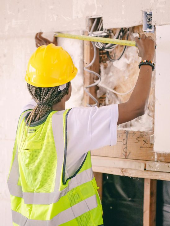 A woman wearing a hard hat and safety vest is measuring a wall with a tape measure.