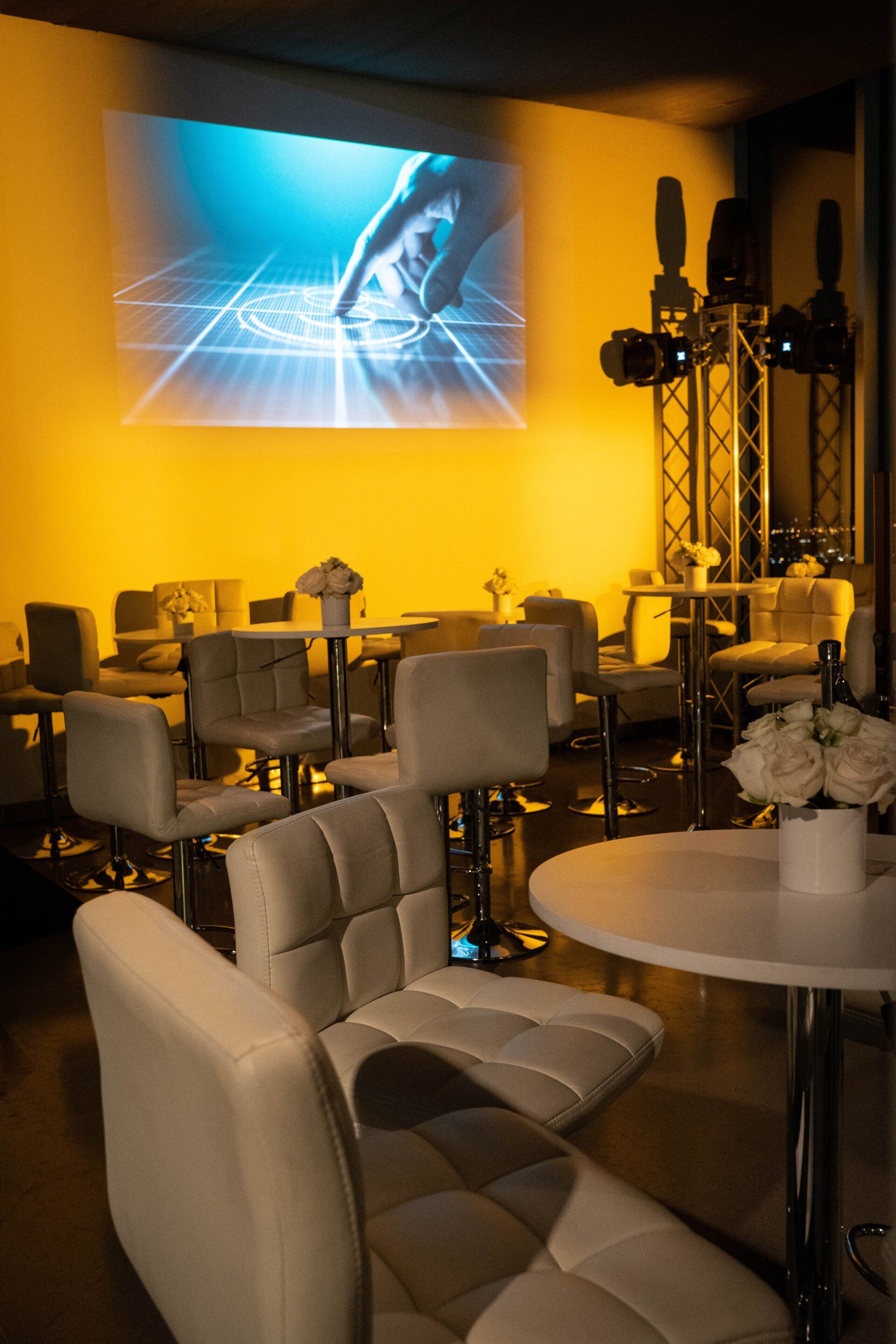 A room with tables and chairs and a projector screen on the wall.