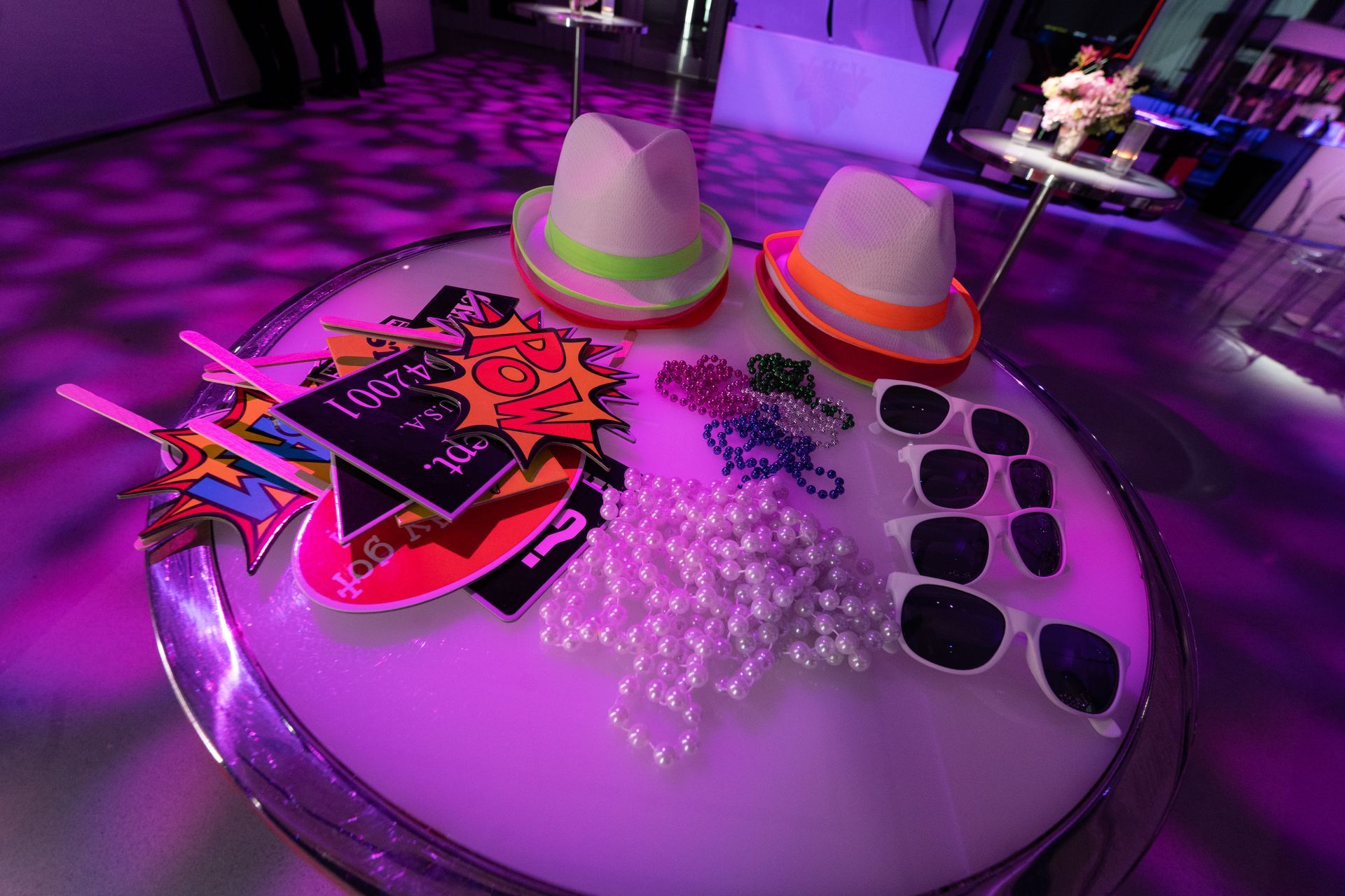 Party photo booth props on a round table: hats, sunglasses, signs, and beads. Purple lighting.
