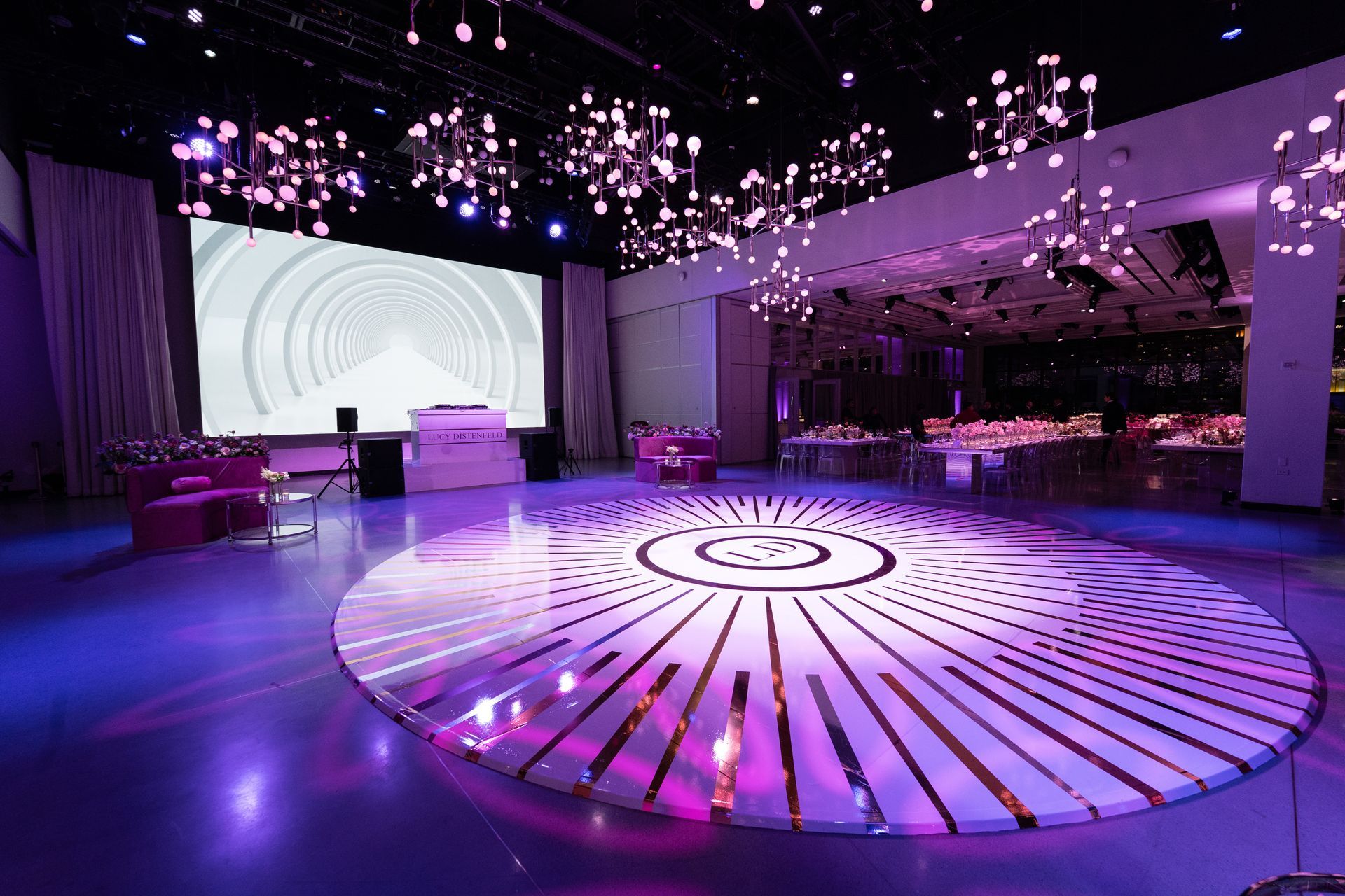 Large event space with illuminated dance floor and stage, purple lighting, decorative chandeliers.