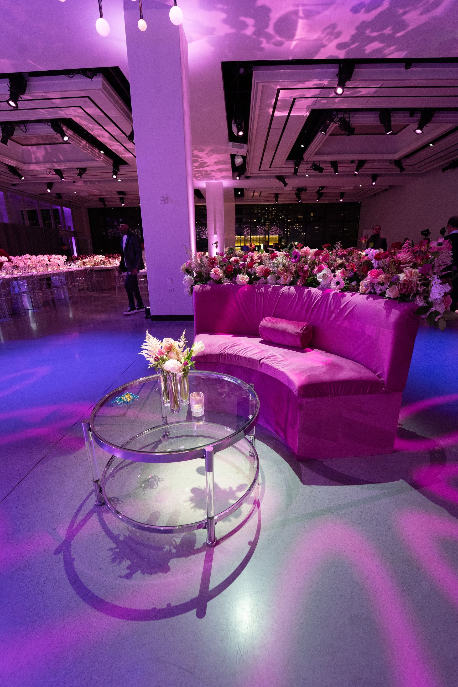 Pink velvet sofa with floral arrangement in a brightly lit room. Glass coffee table, purple lighting.