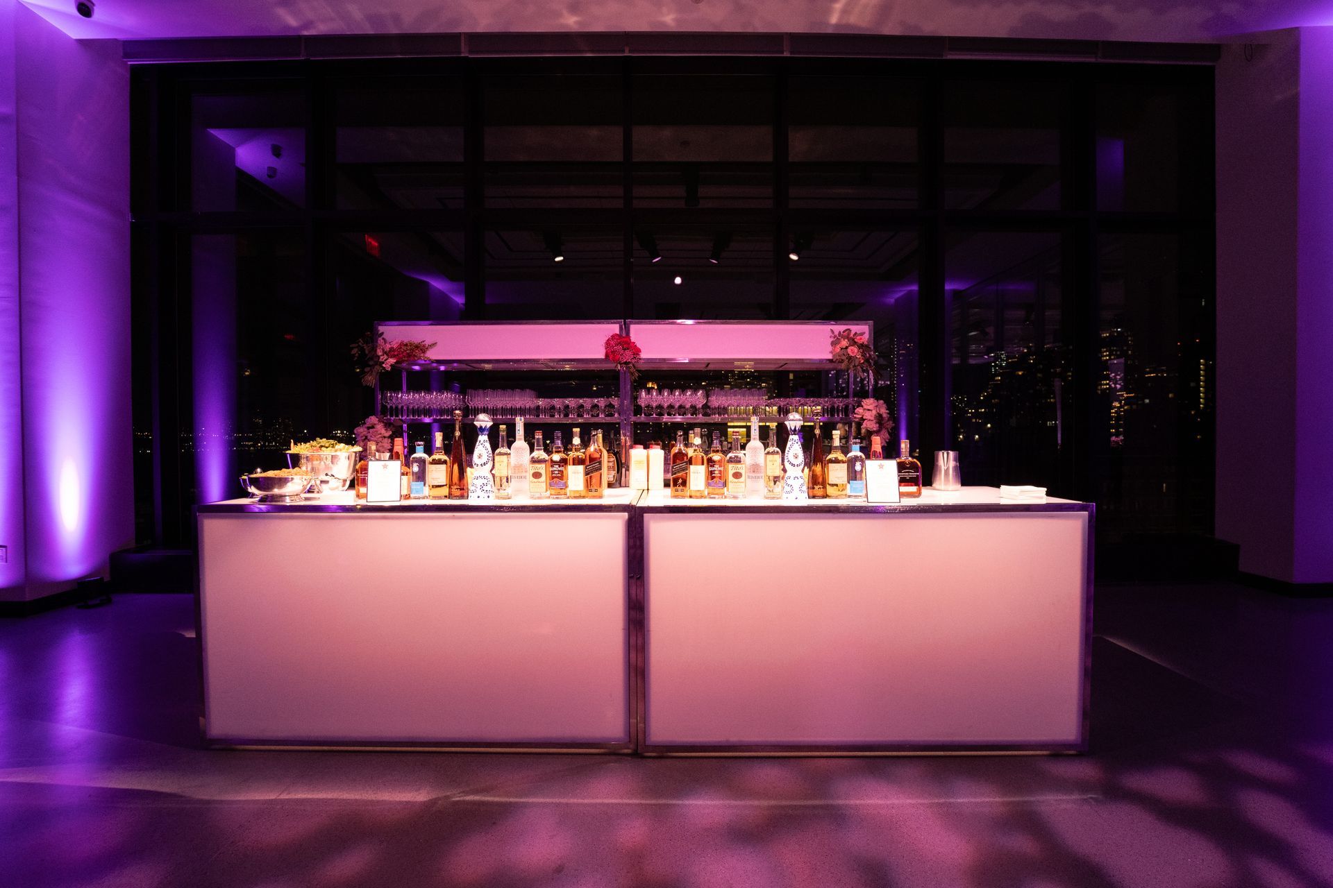 A lit bar with alcohol bottles, glasses, and a bartender, set against a dark window and purple lighting.