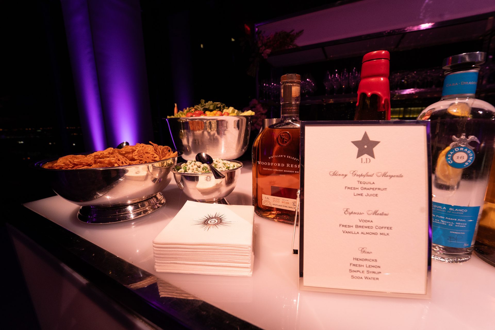 Bar setup with bowls of food and bottles, a menu, and napkins under purple lighting.