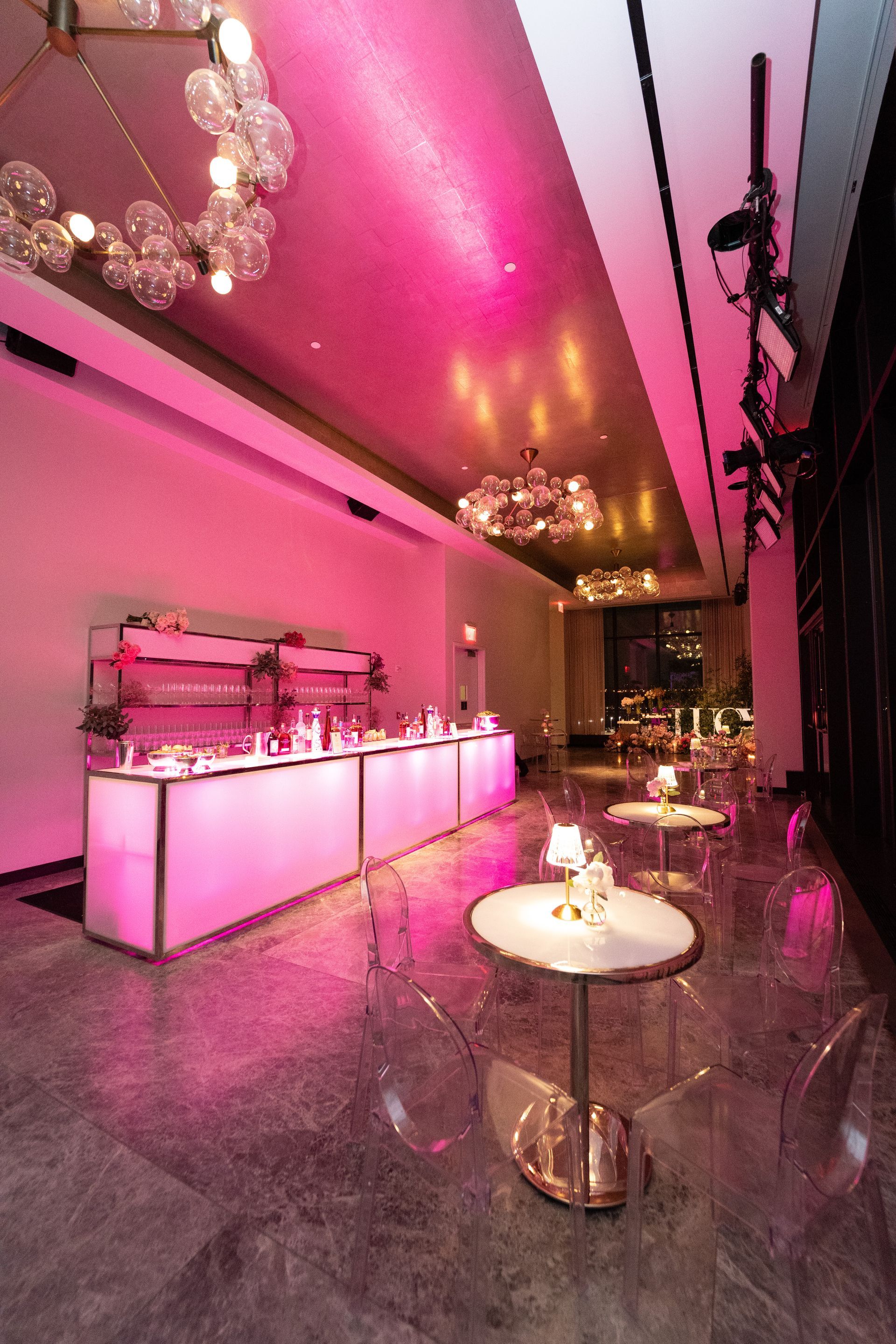 A bar area with pink lighting, tables, and transparent chairs. Two globe chandeliers hang from the ceiling.