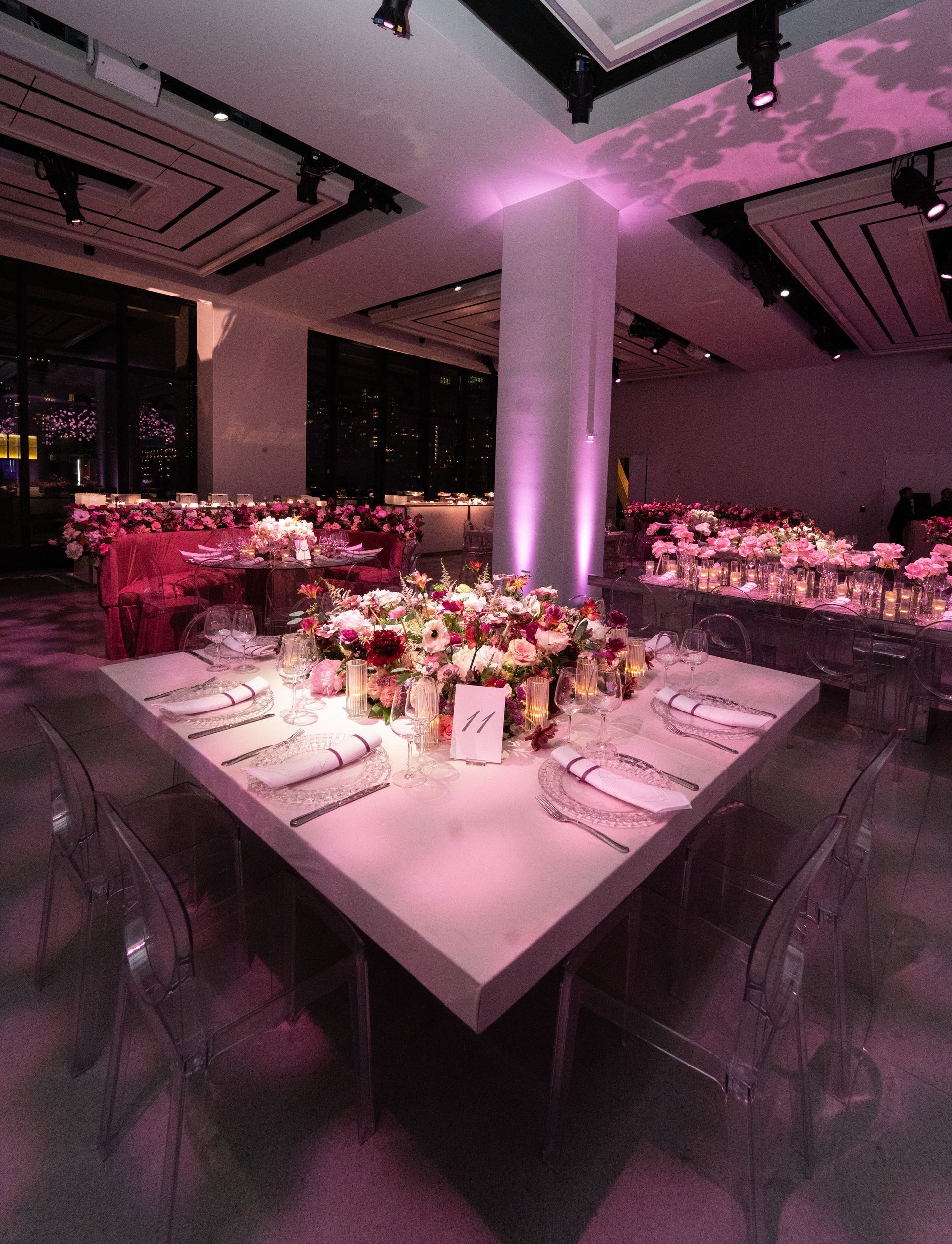 Elegant dining hall with a large square table, flowers, and pink lighting.
