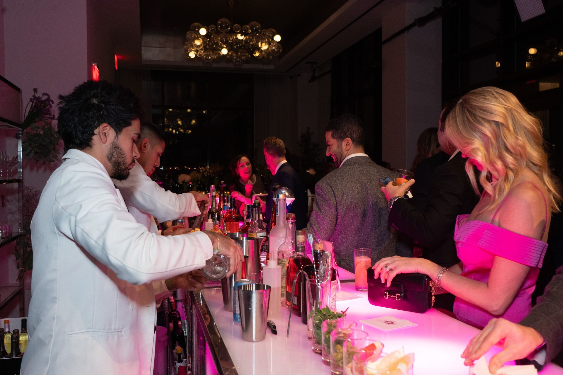Bartender mixing drinks at a brightly lit bar. People mingle, a woman in pink dress reaches for a drink.