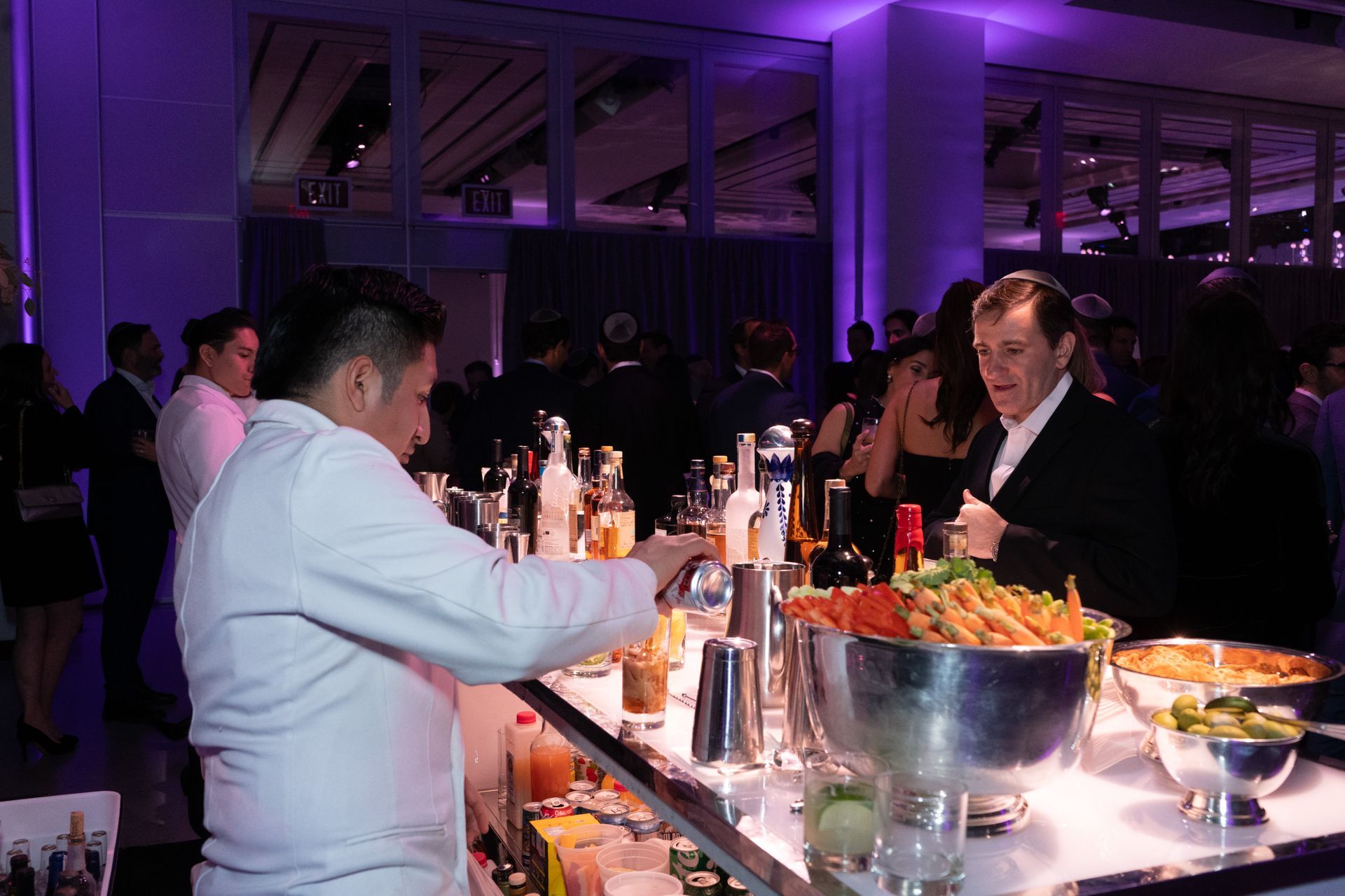 Bartender mixing drinks at a brightly lit bar during an event. People in formal attire in the background.