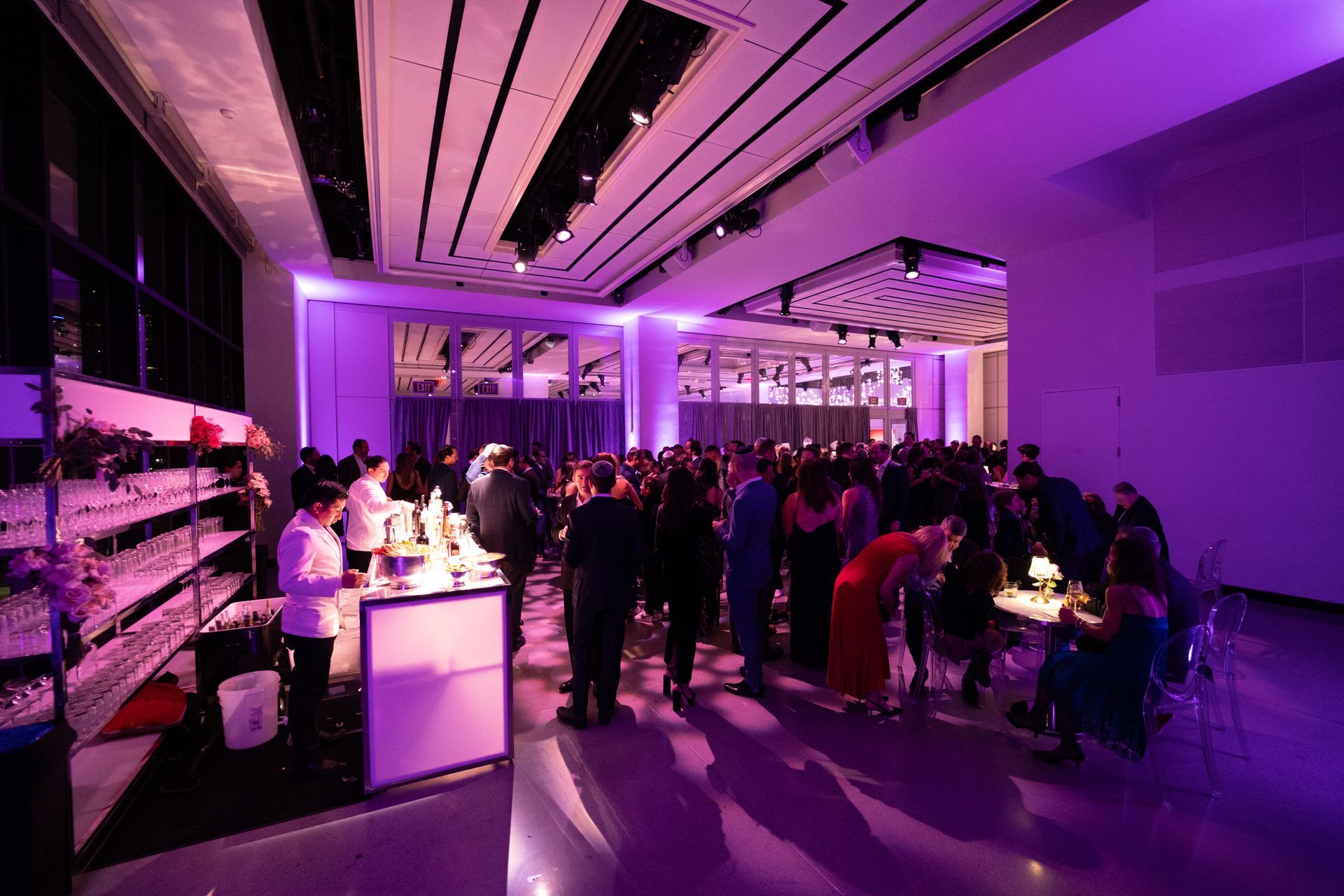Party in a modern hall lit with purple. People gather near a bar, enjoying an event.