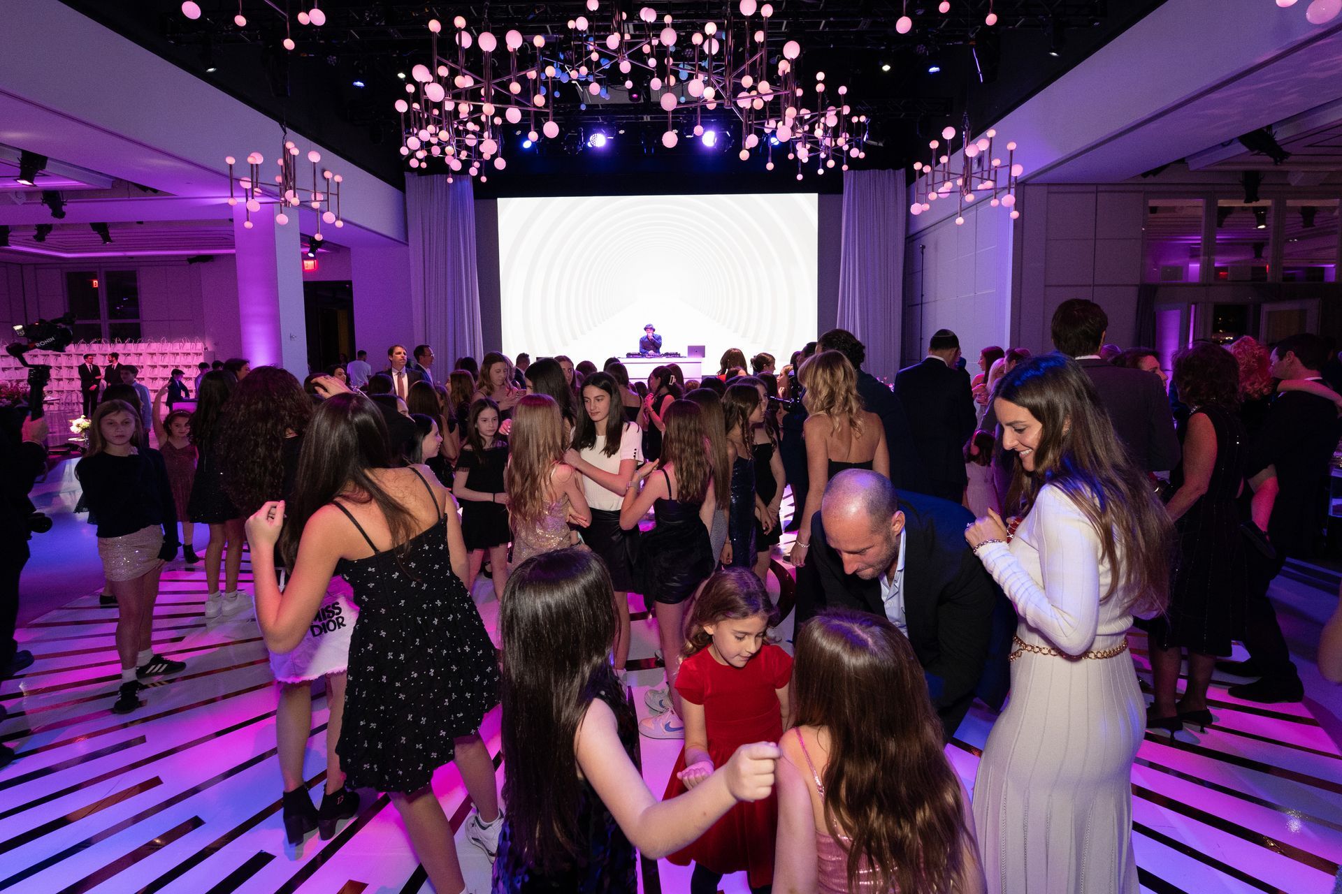 People dancing at a party with a light-up dance floor, DJ in the background, and decorative ceiling lights.