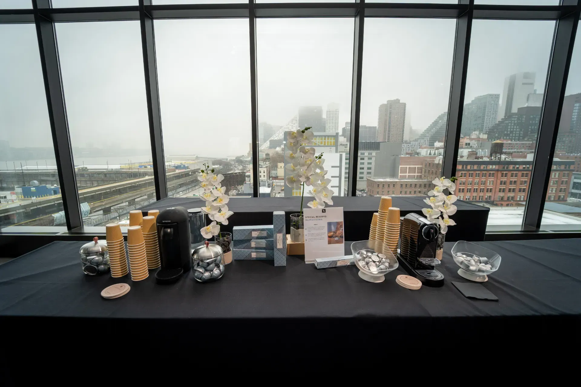 A table with candles and flowers on it in front of a window with a city in the background.