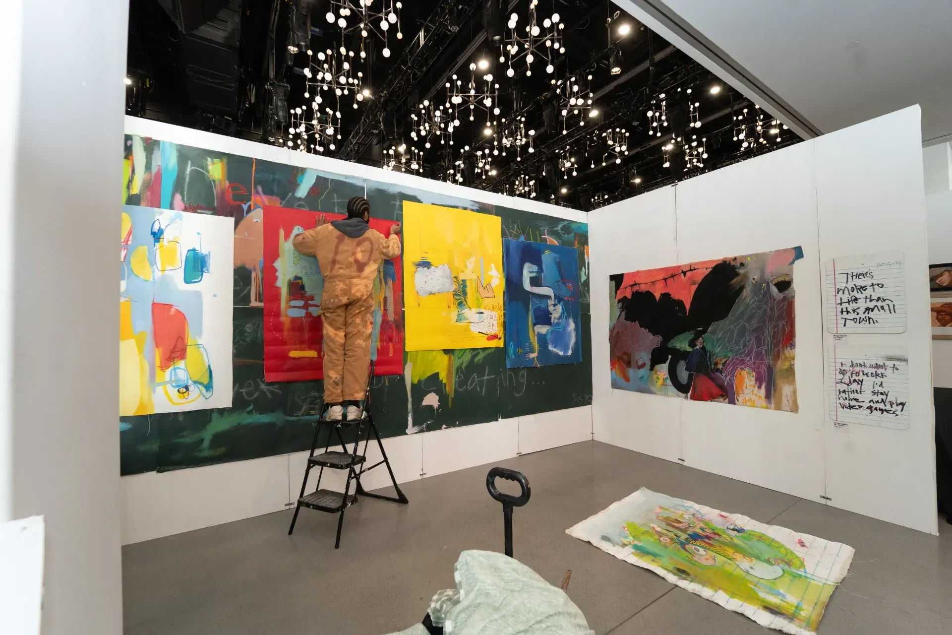 A man is standing on a ladder in front of a wall with paintings on it.