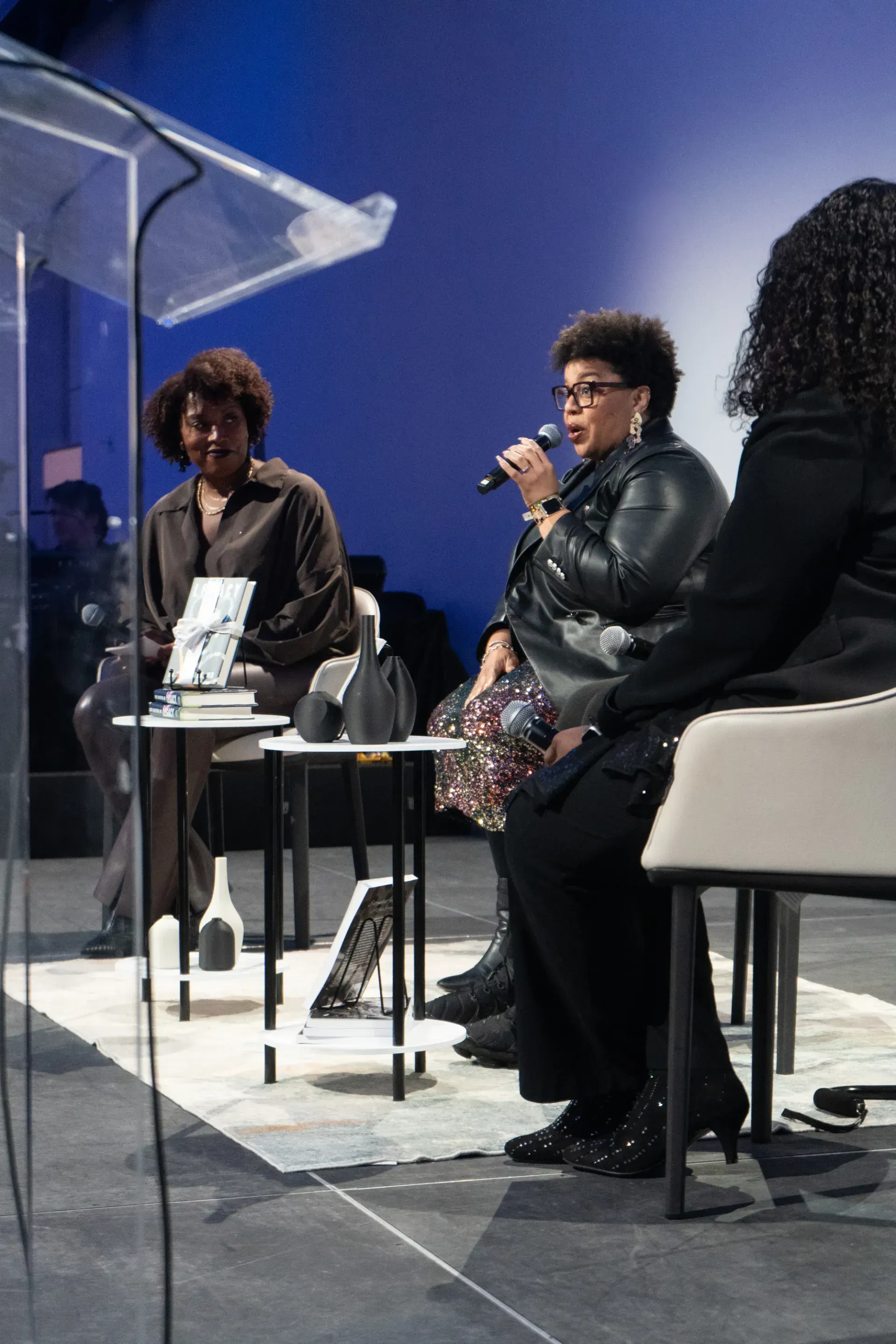 A woman is speaking into a microphone while two other women sit at tables.
