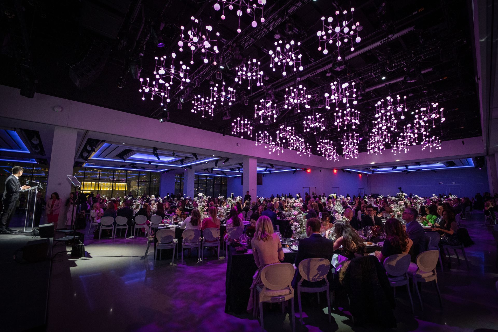 Large event in a hall, tables set with guests. Purple lights hang from the ceiling. A speaker on a stage.
