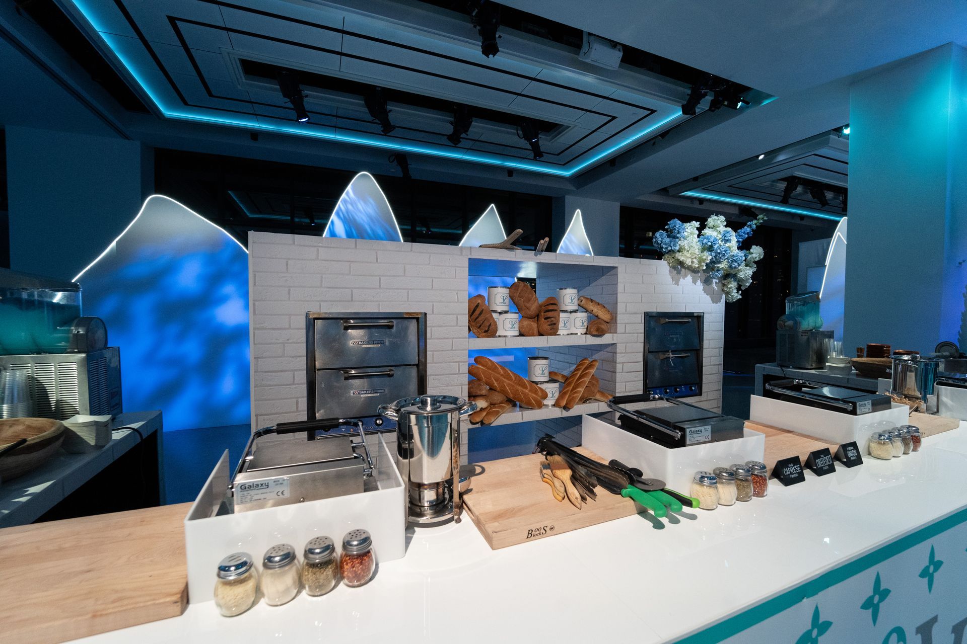 Food service counter with bagels and various kitchen equipment, bathed in blue light.