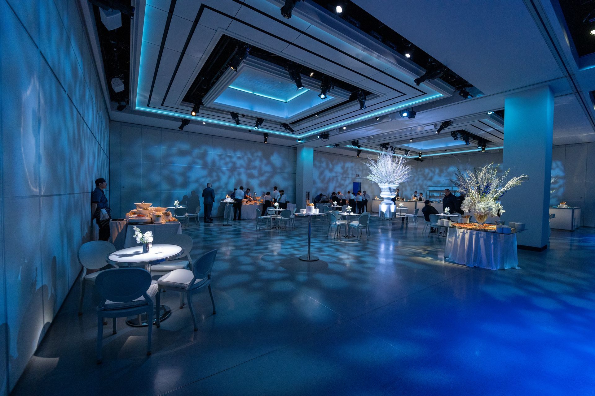 An event hall lit in blue. Guests mingle near tables with food and floral arrangements.