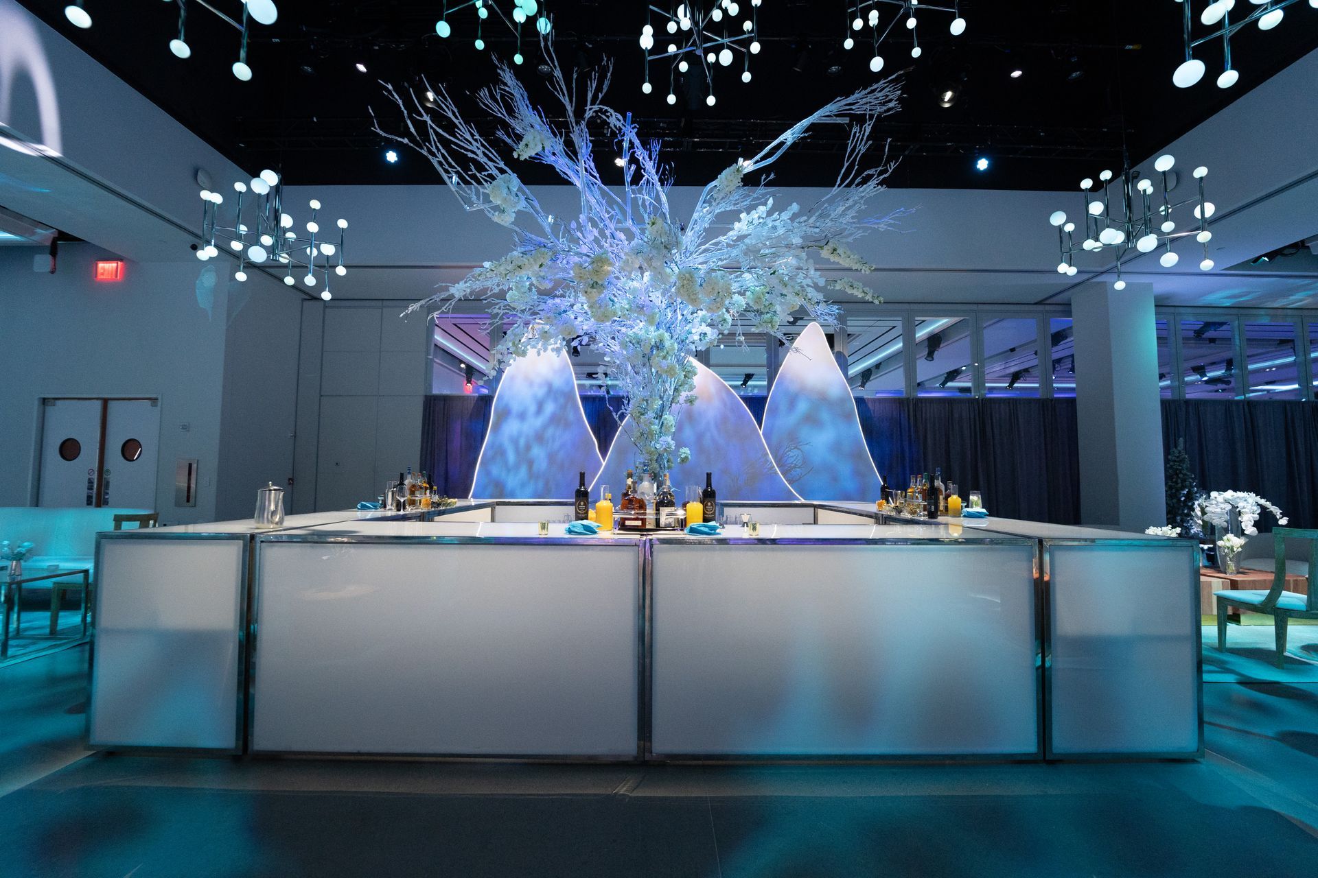 A bar with white panels, bottles, and floral arrangement. A festive indoor event in blue lighting.