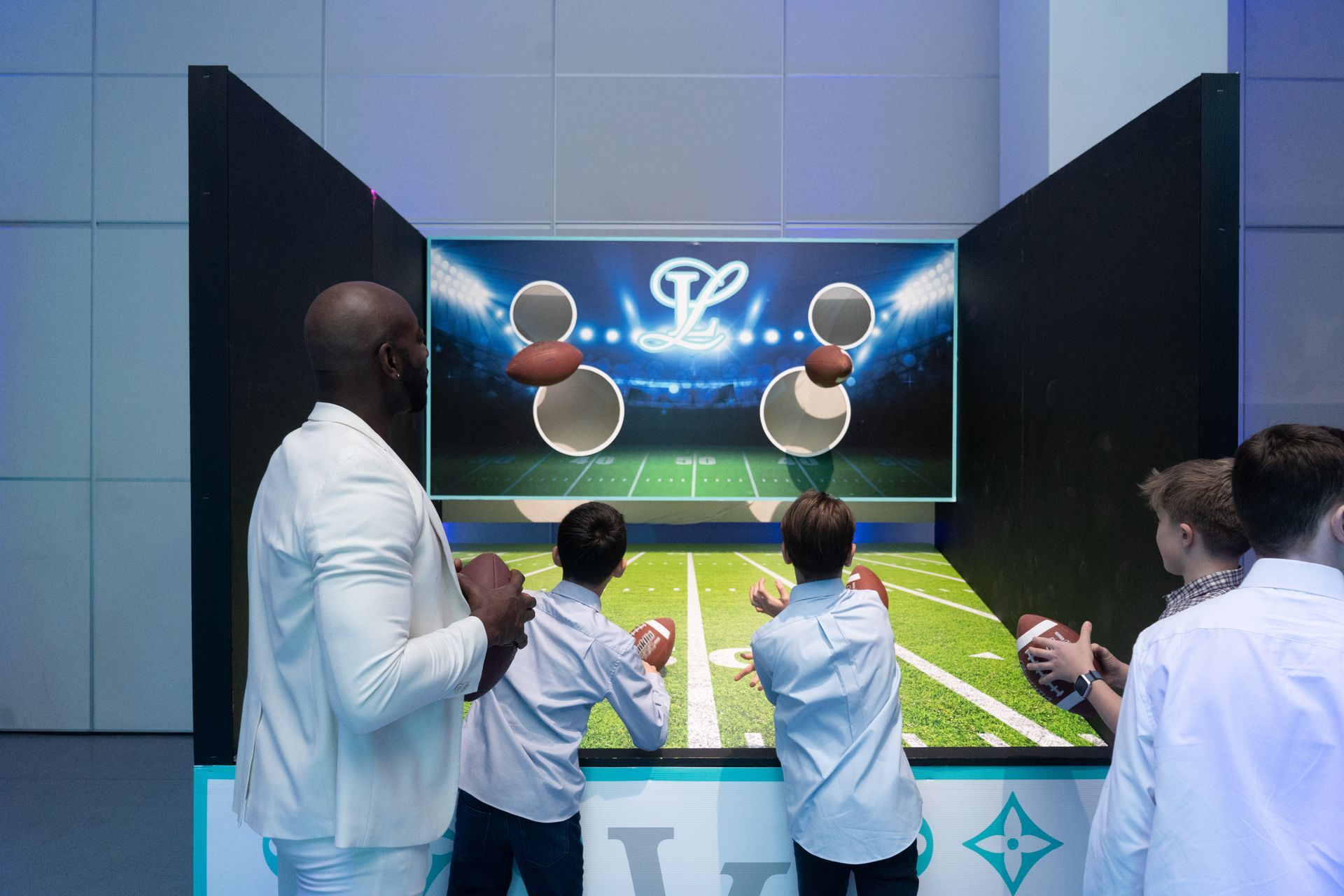 A man in a white suit watches children throwing footballs at a game.