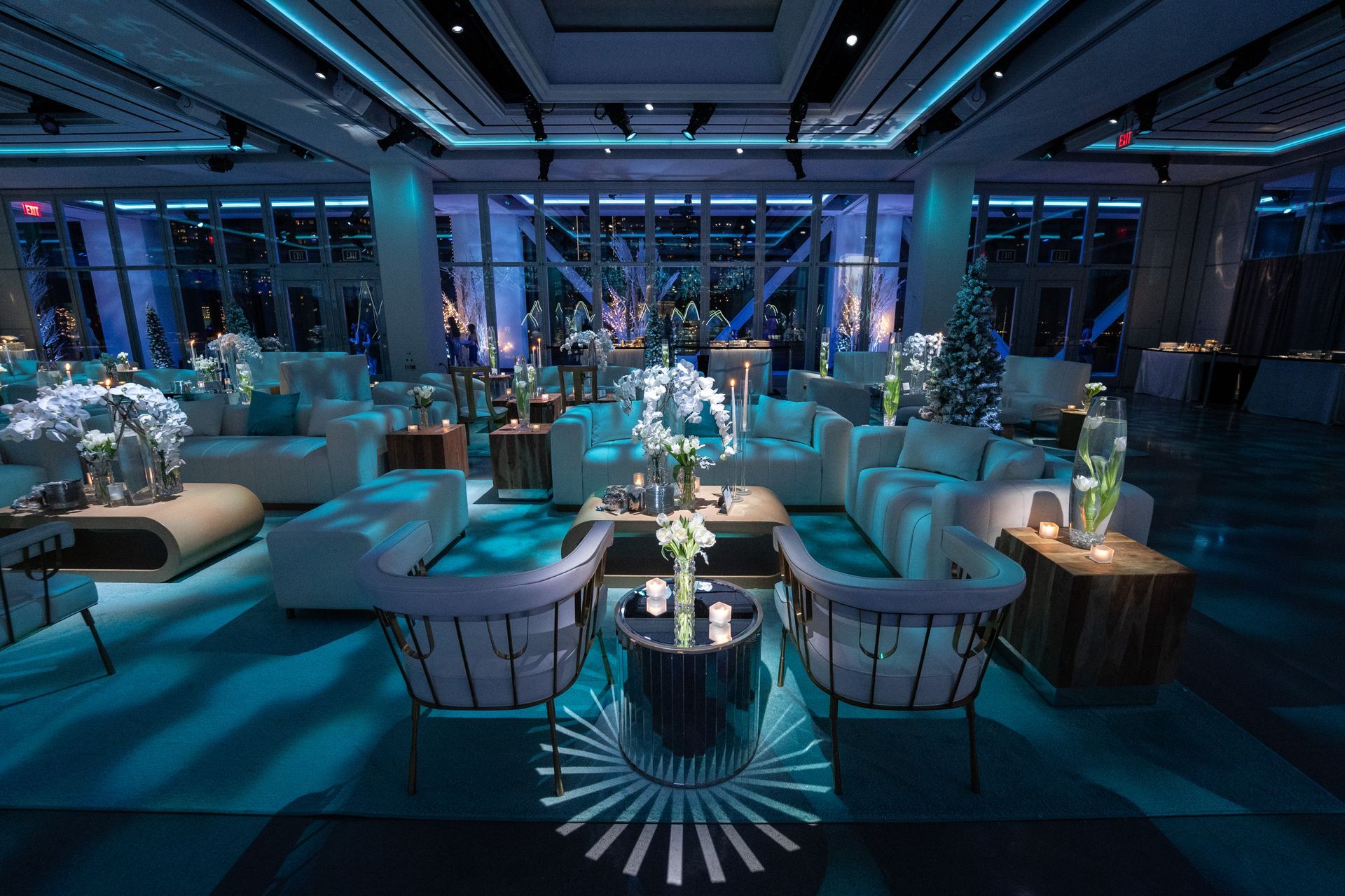 Blue-lit modern lounge with white couches, floral centerpieces, and city views.