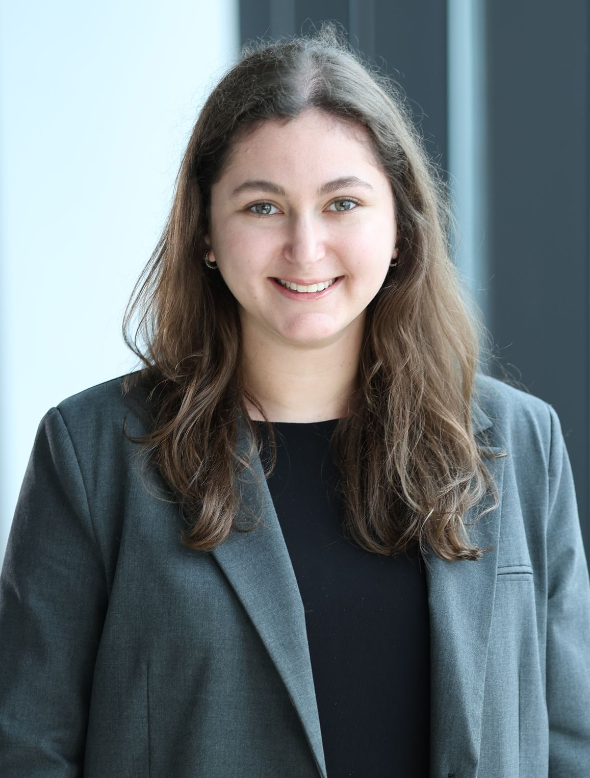 Woman in a grey blazer smiling, standing in front of a window.