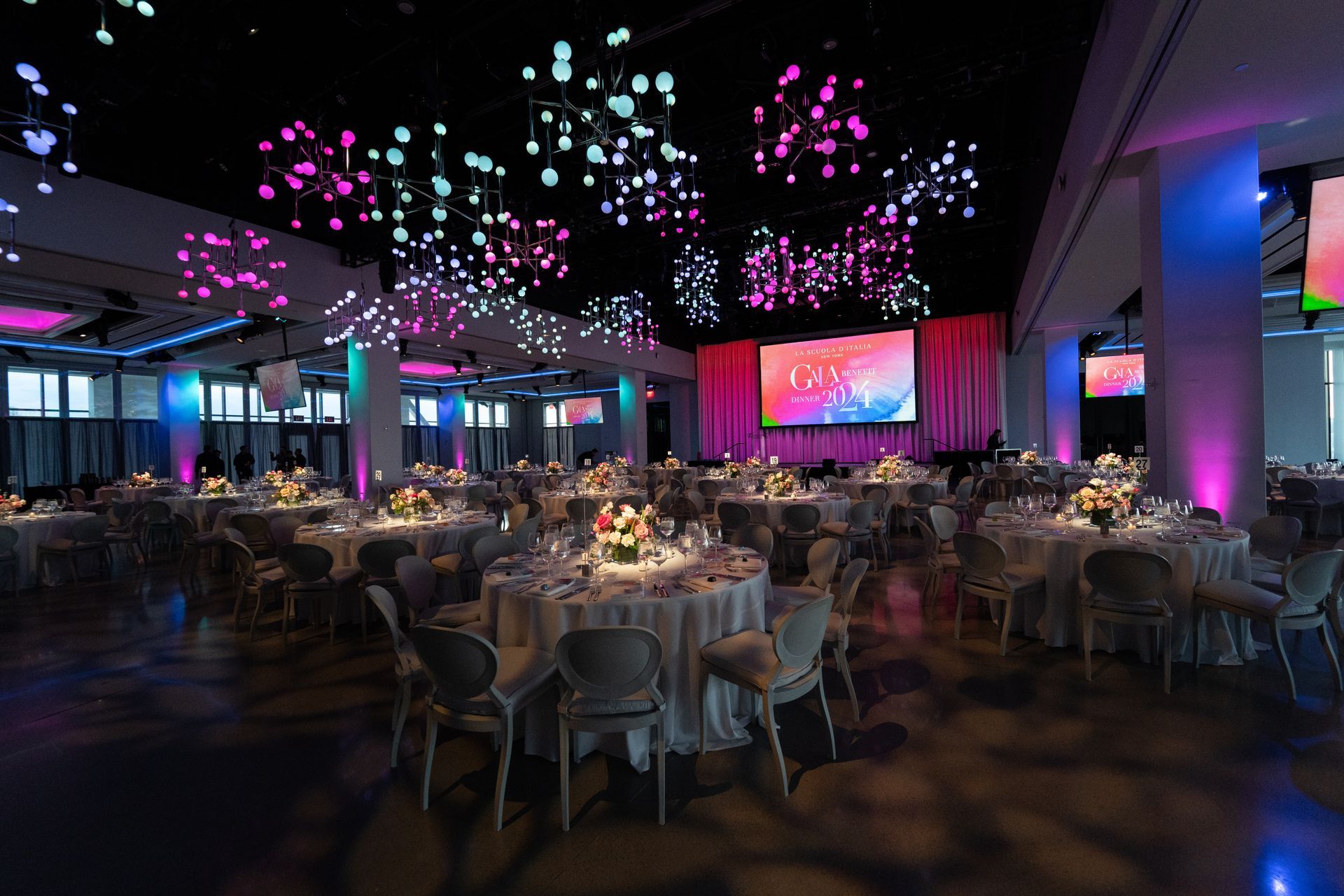 Elegant ballroom with round tables set for a formal event. Overhead, decorative lights and screens display graphics; blue, pink lighting.