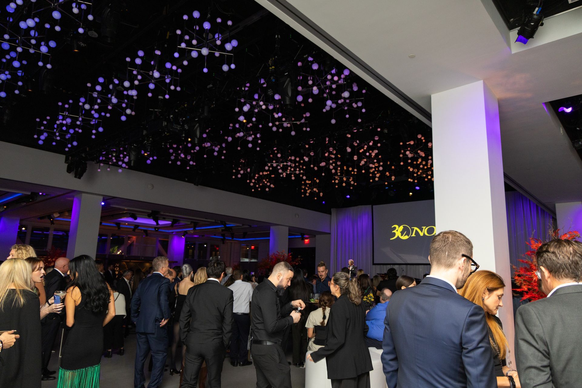 People gather at an event in a room with a decorated ceiling. Purple and gold lights.
