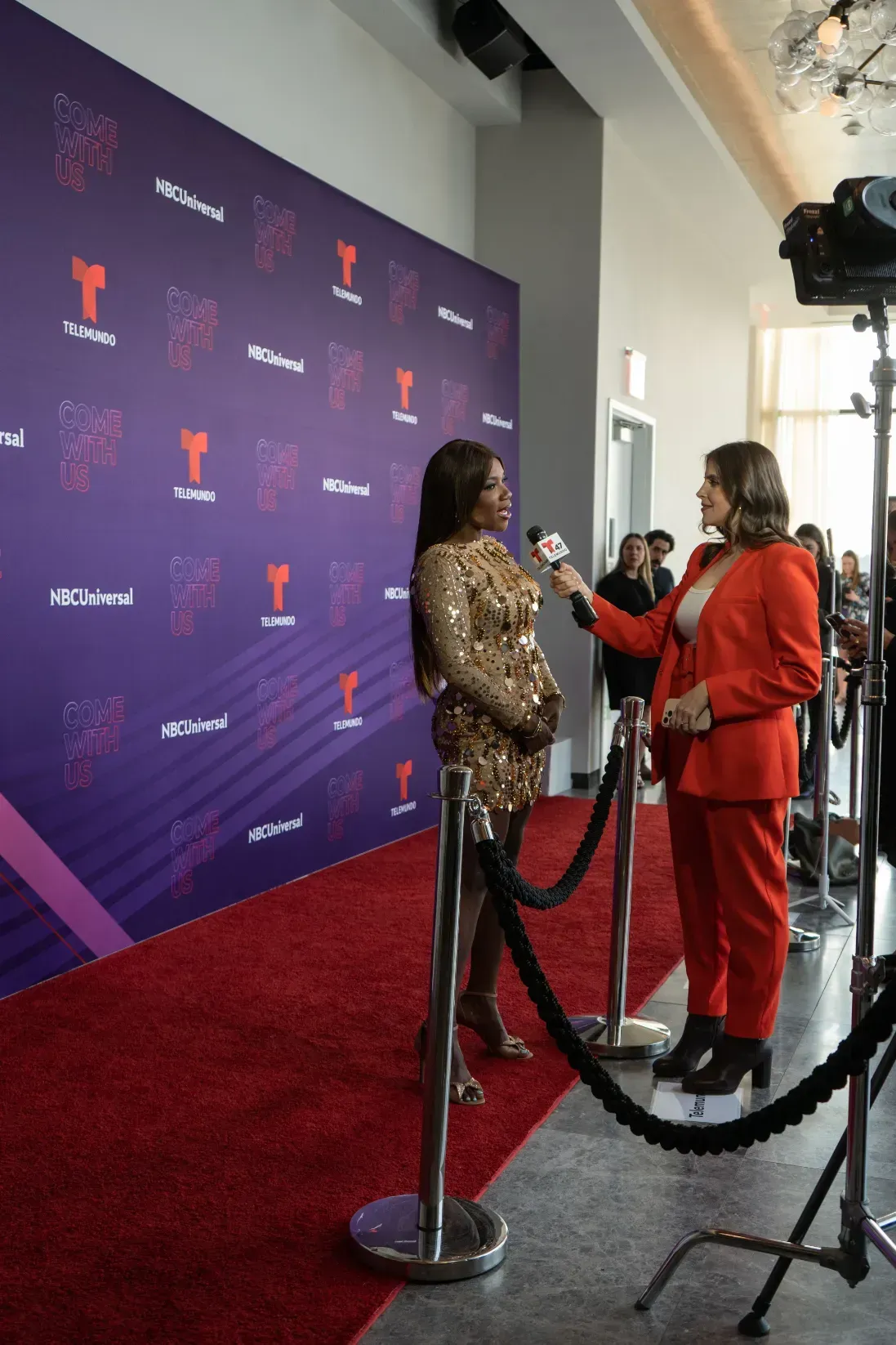 A woman in a red suit is talking to a woman on a red carpet.