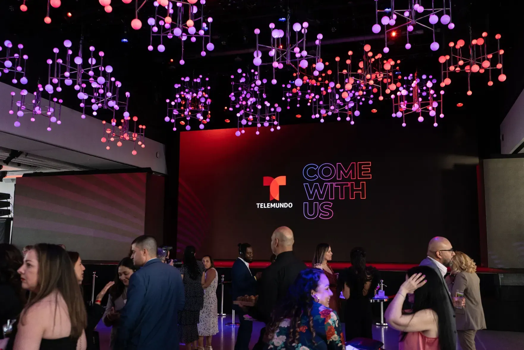 A group of people are standing in a room with a large screen that says come with us.