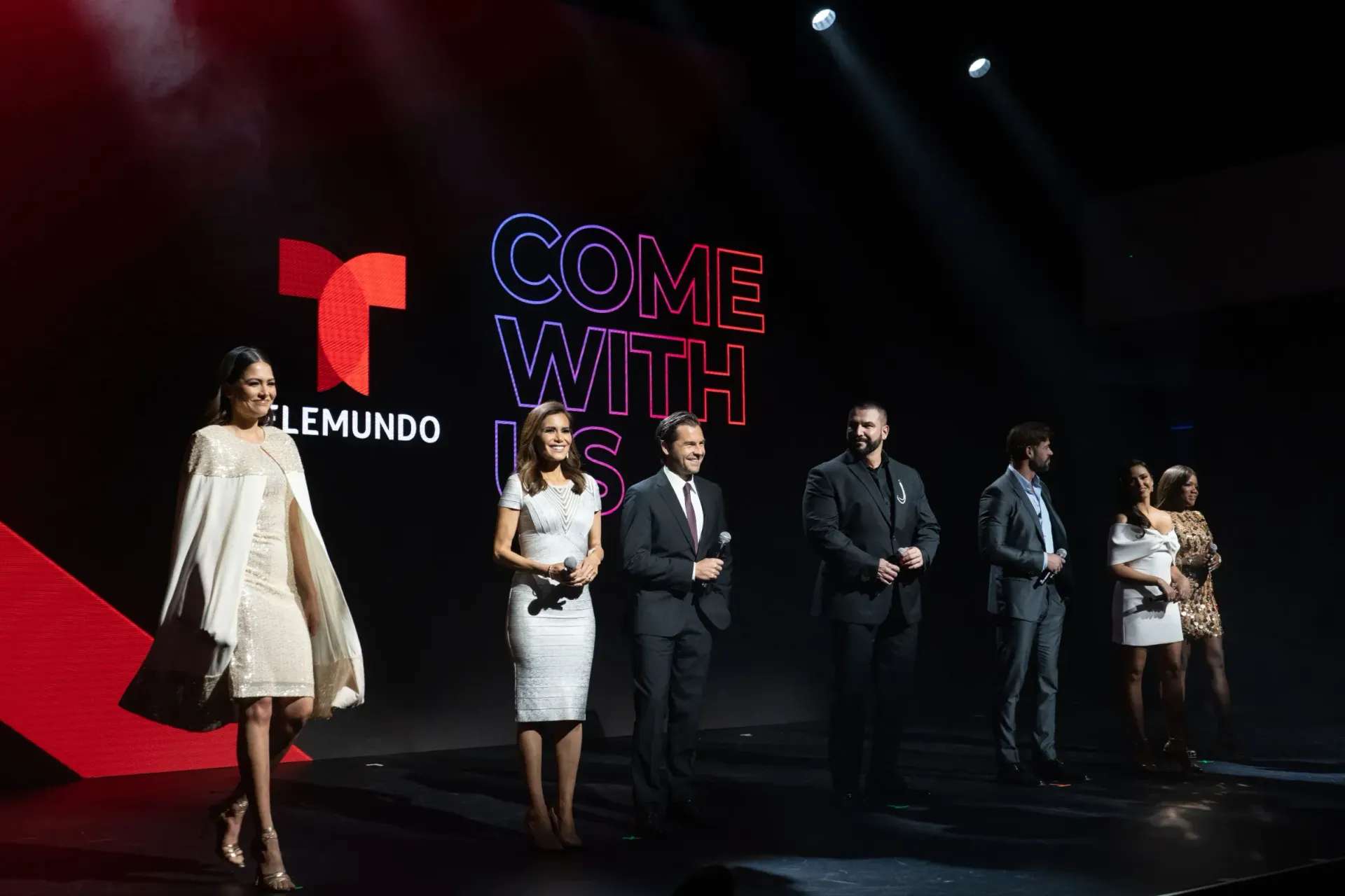 A group of people are standing on a stage in front of a sign that says come with us.