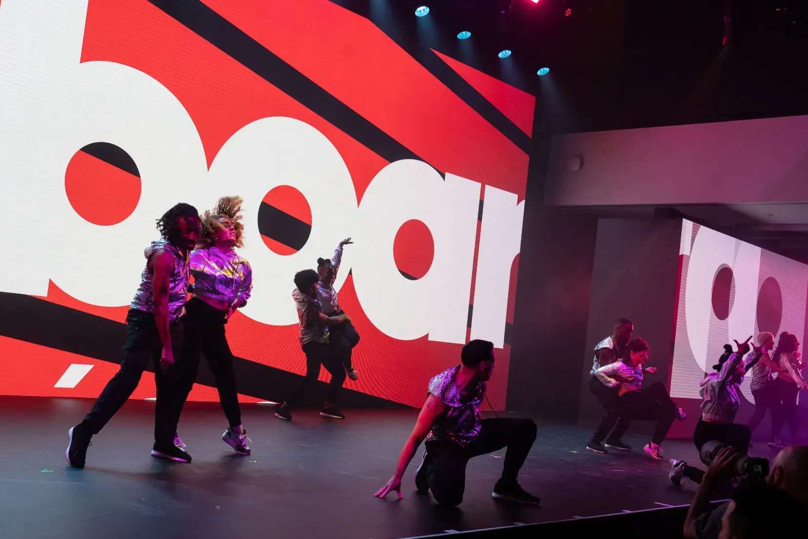 A group of people are dancing on a stage in front of a large screen with the word dear on it.