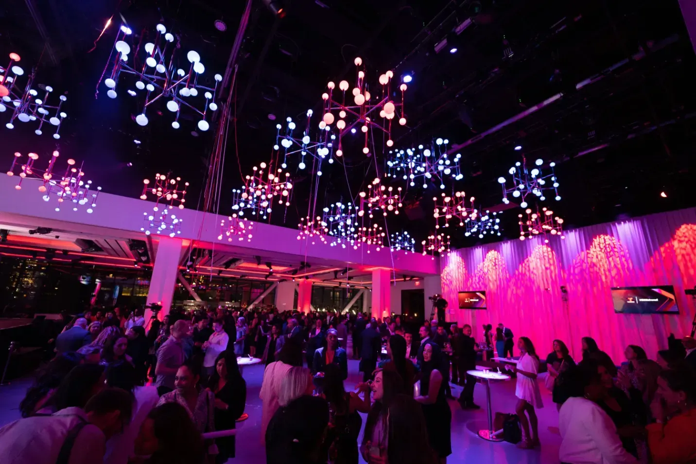 A crowd of people are dancing in a large room with purple lights on the ceiling.