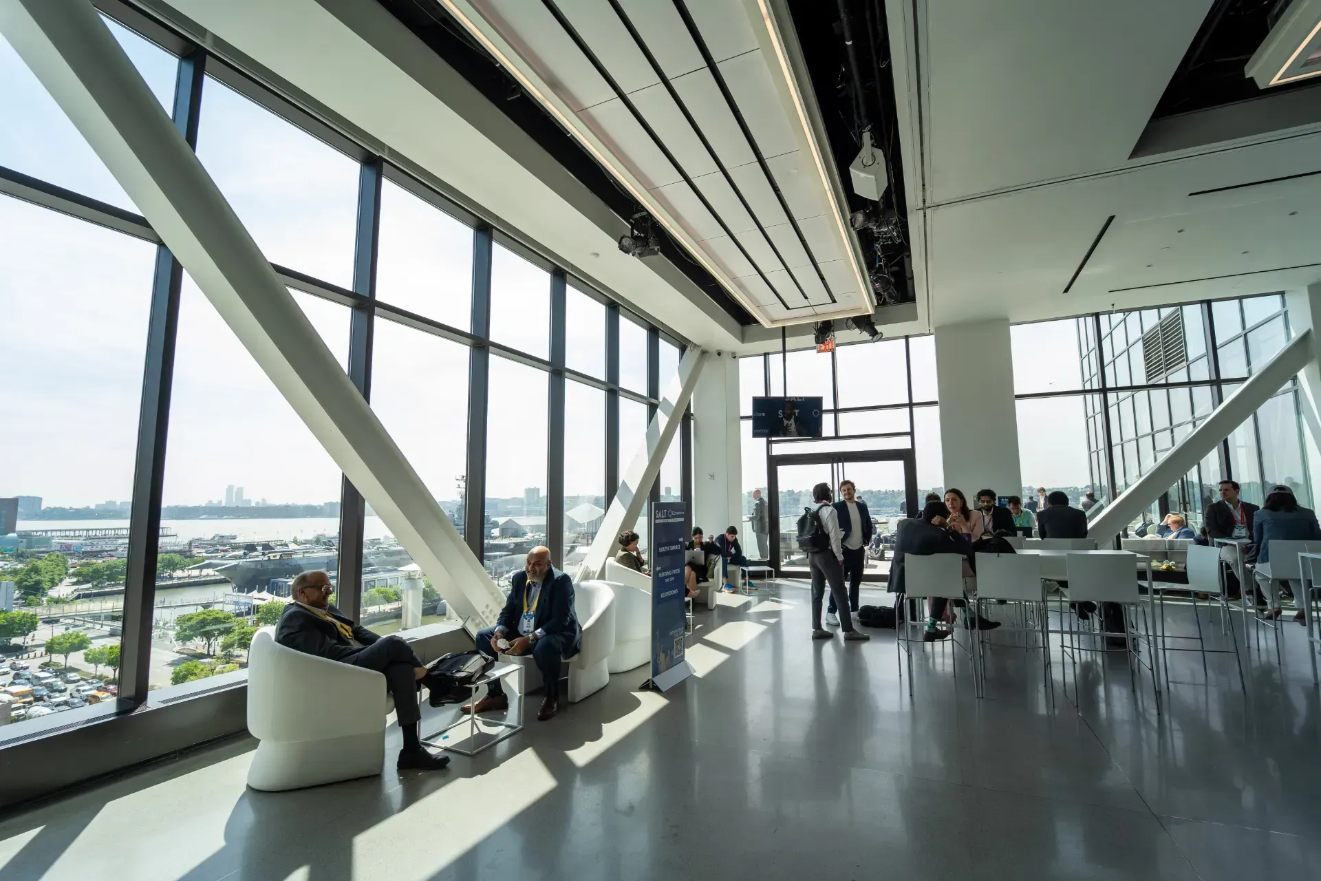 A group of people are sitting in chairs in a large room with a lot of windows.