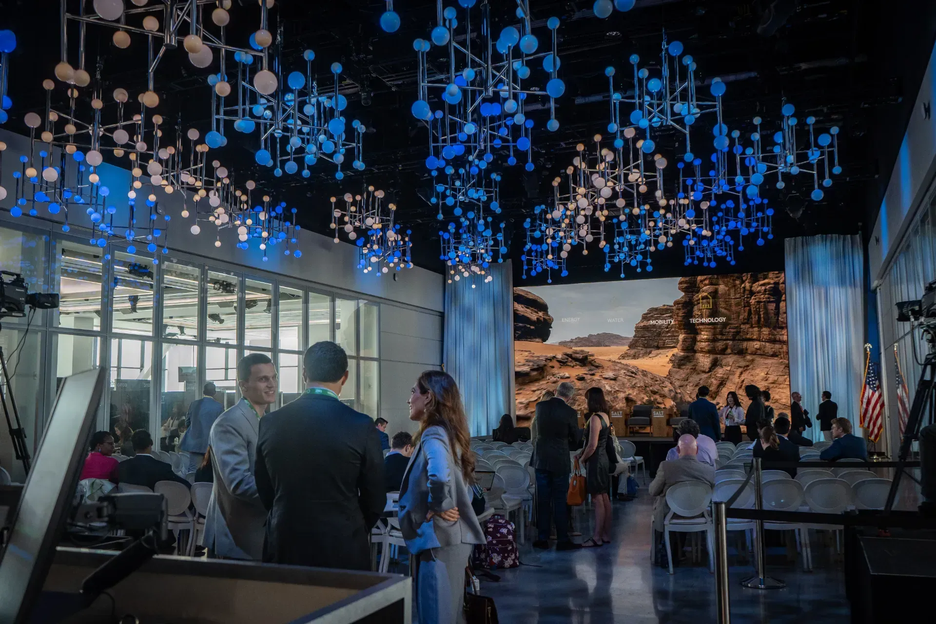 A group of people are standing in a room with a large screen hanging from the ceiling.