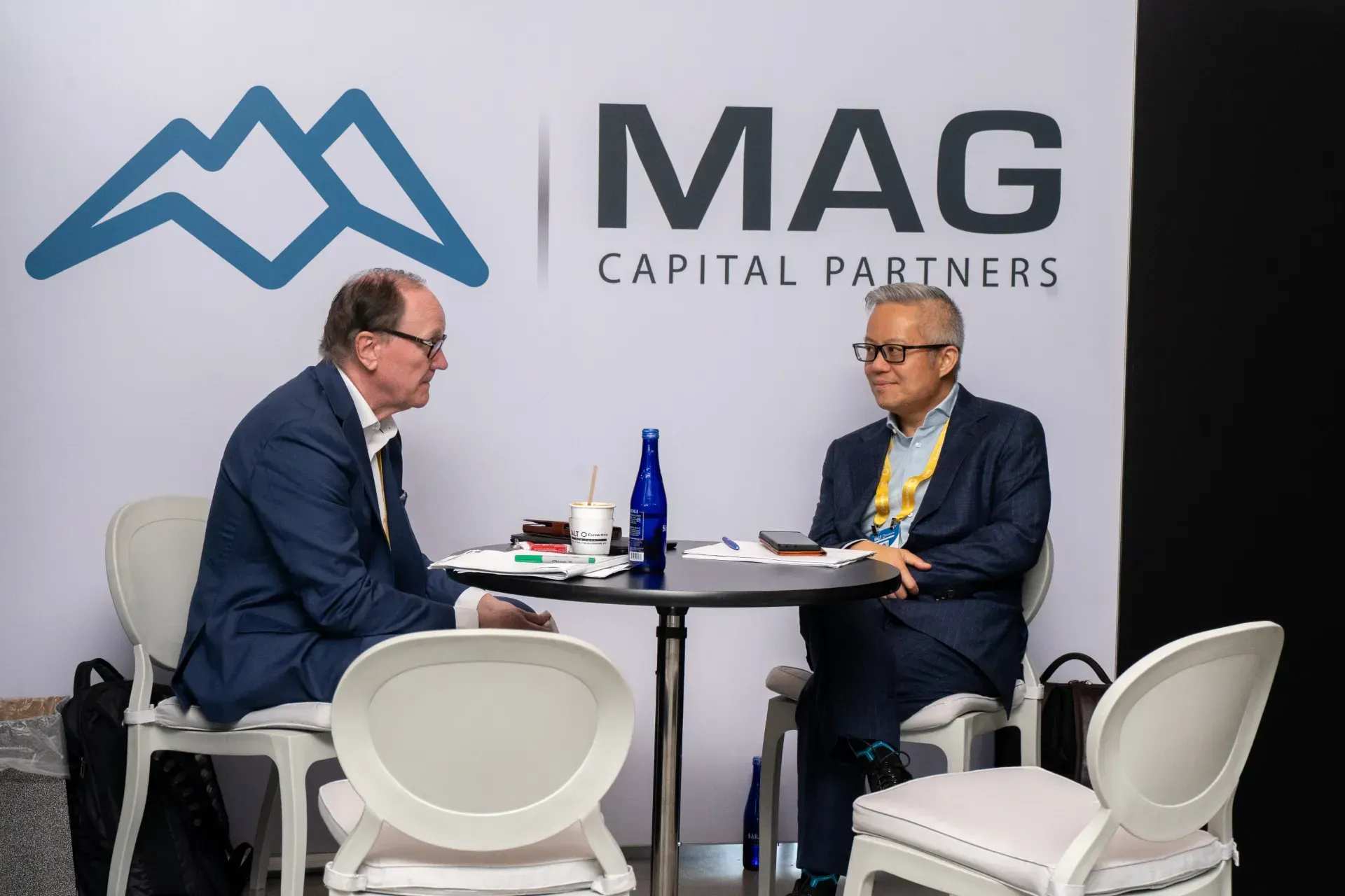 Two men are sitting at a table in front of a mag capital partners sign.