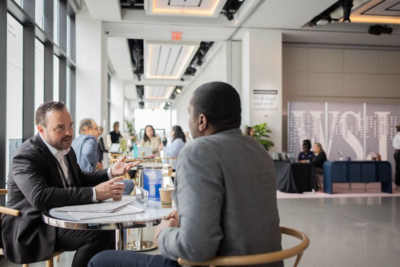 Two men seated at a table converse in a bright, open space, possibly at an event.