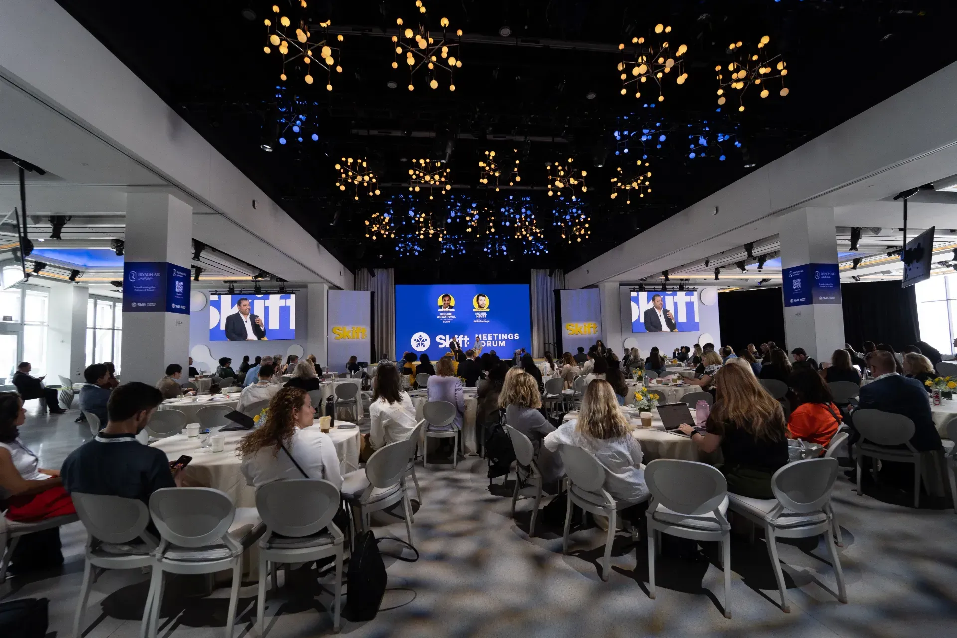 A group of people are sitting at tables in front of a large screen.