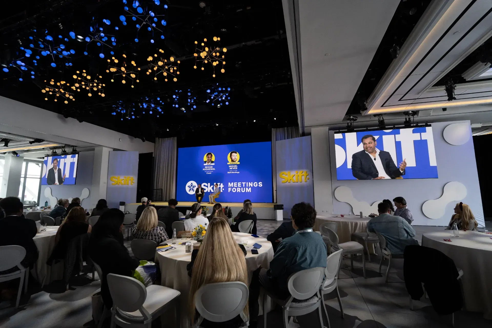 A group of people are sitting at tables in front of a large screen.