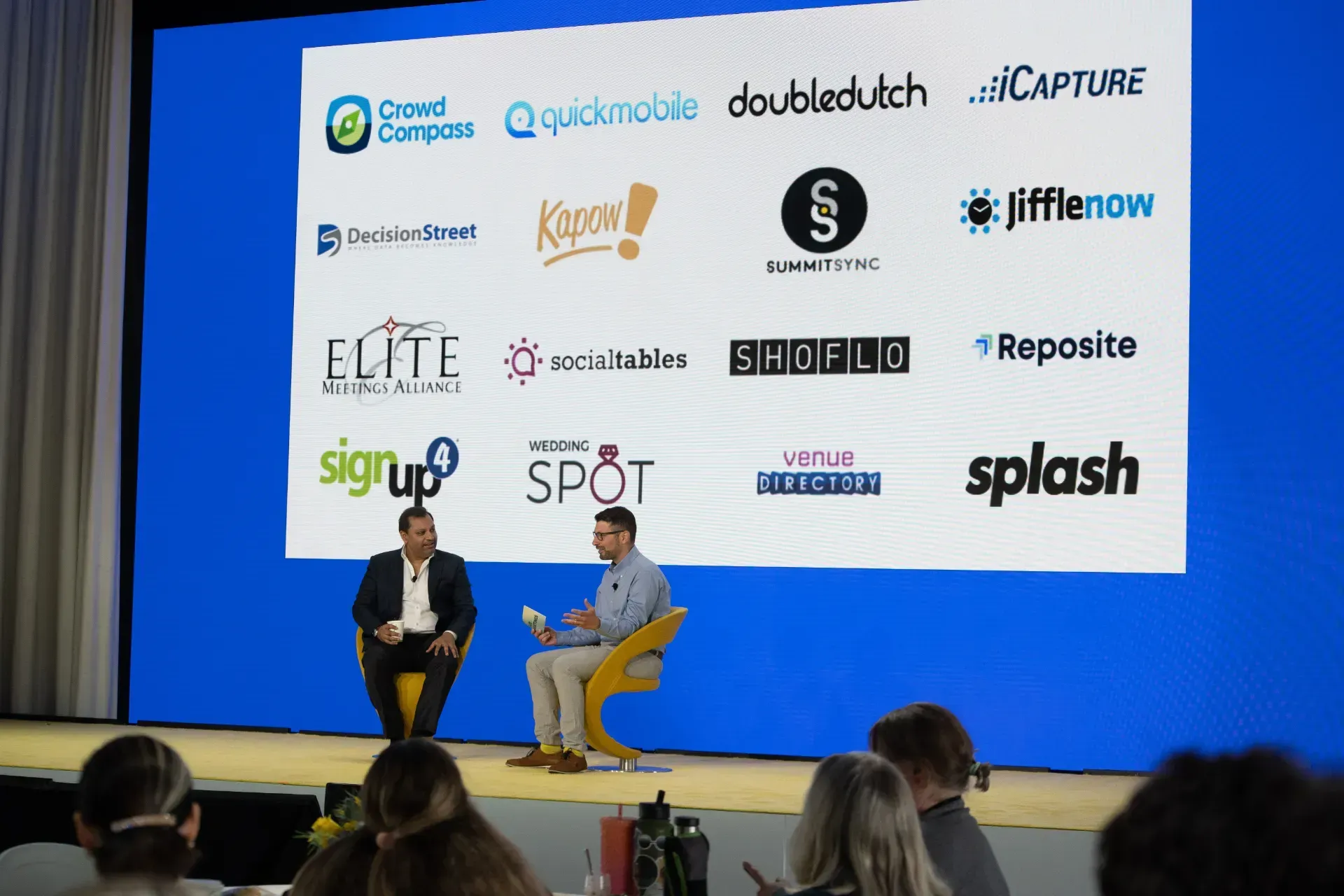 Two men are sitting on a stage in front of a large screen with logos on it.
