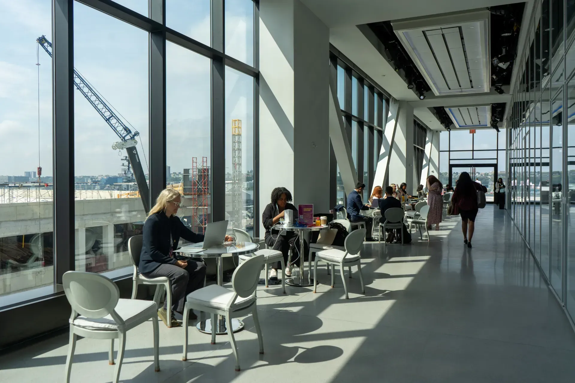 A group of people are sitting at tables in a large room with lots of windows.