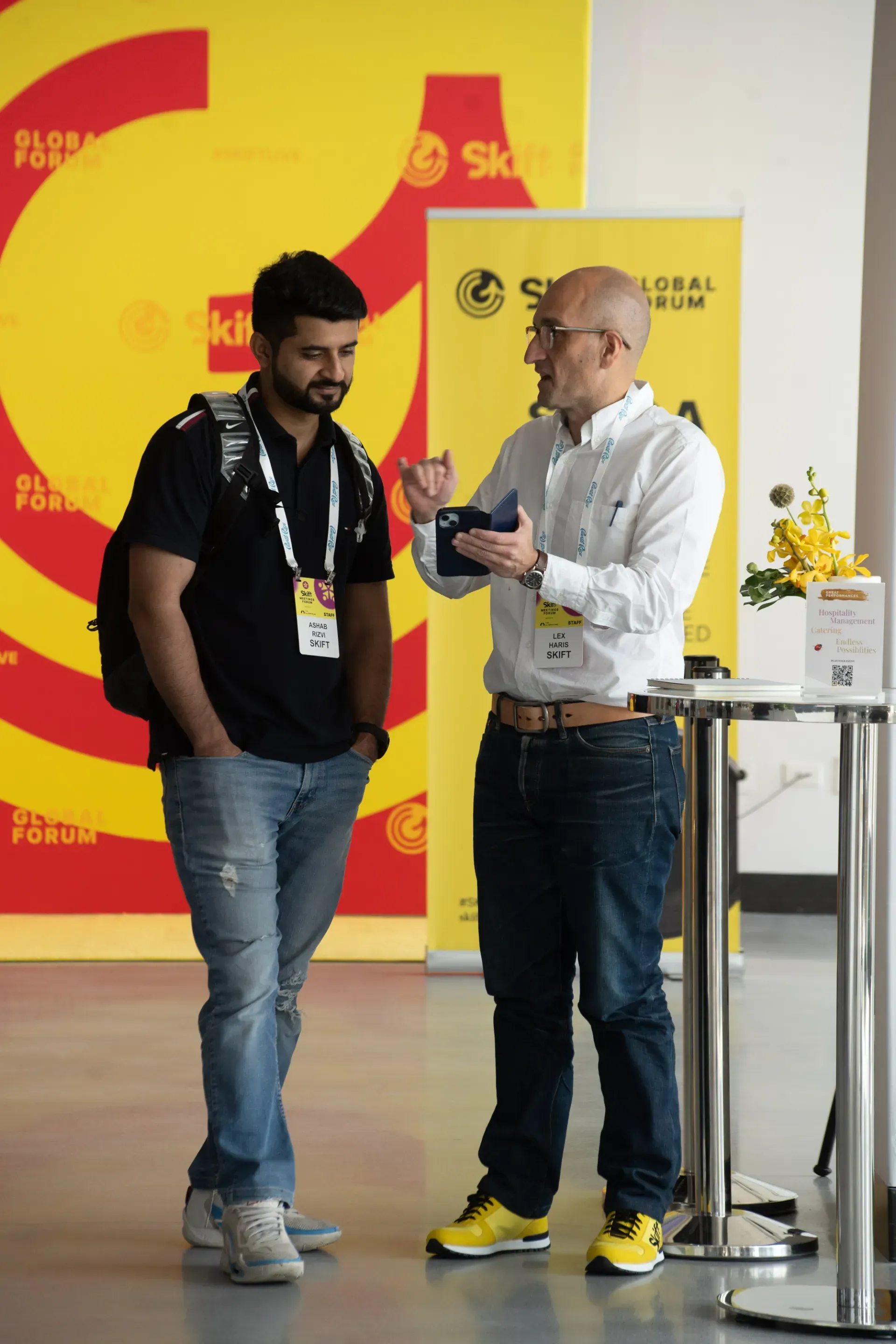 Two men are standing next to each other in front of a yellow wall.