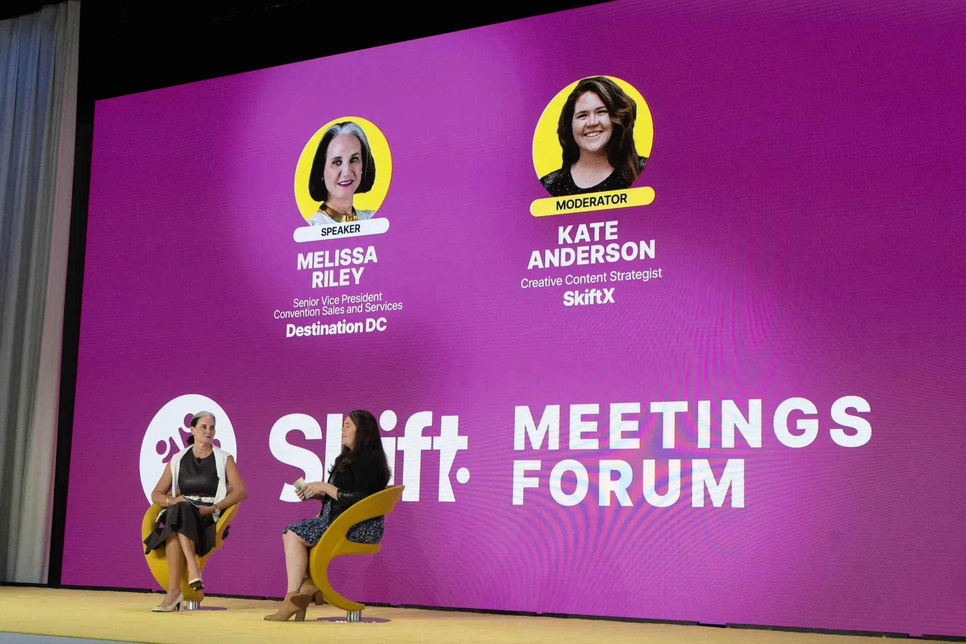 Two women are sitting in front of a large screen that says shift meetings forum.