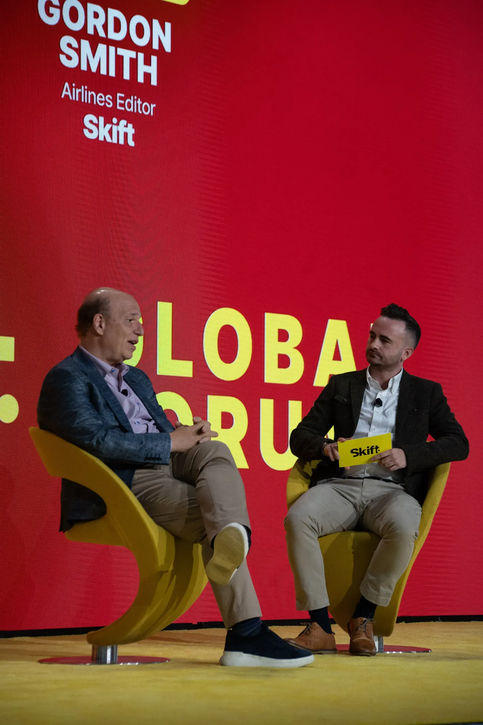 Two men are sitting in chairs on a stage talking to each other.
