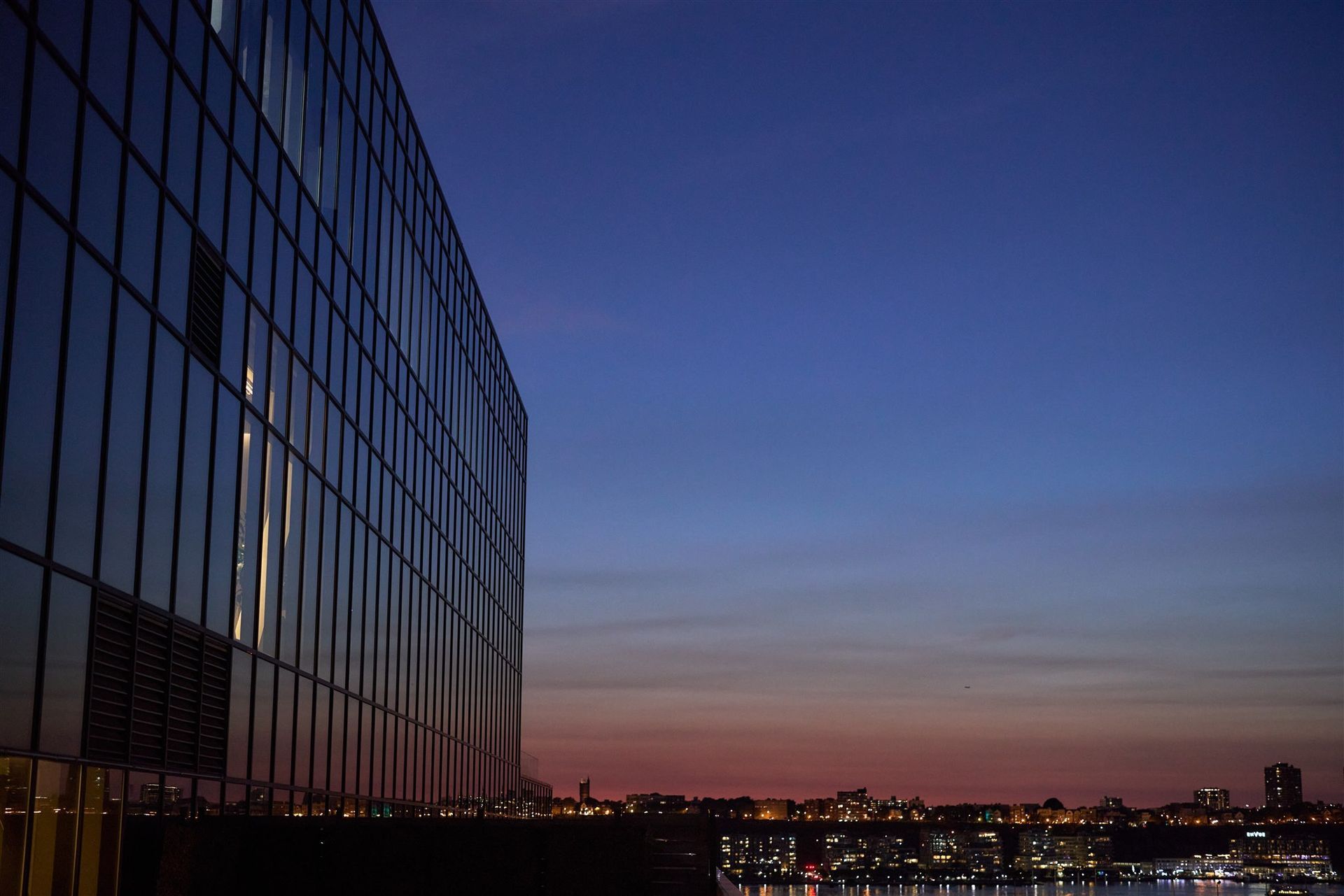 A rooftop patio at sunset with dark lounge seating, a central coffee table, and city views near a glass building.